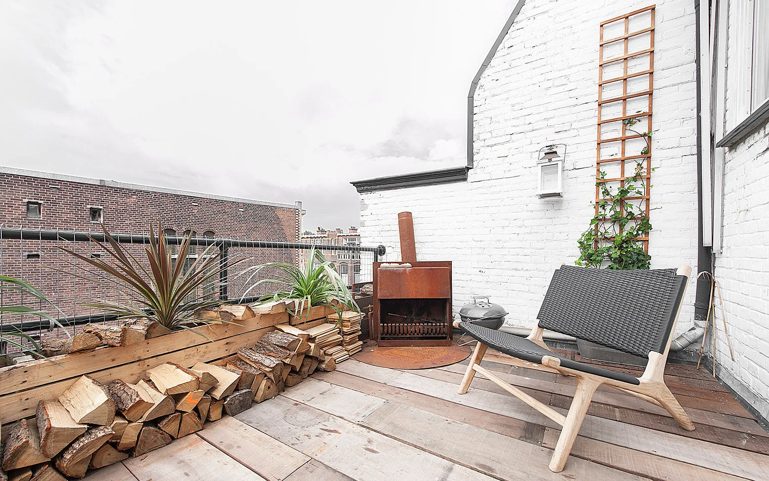 A rooftop patio features a woven lounge chair, a rusted metal fire pit, and stacked firewood beneath a wooden planter against a white brick wall.