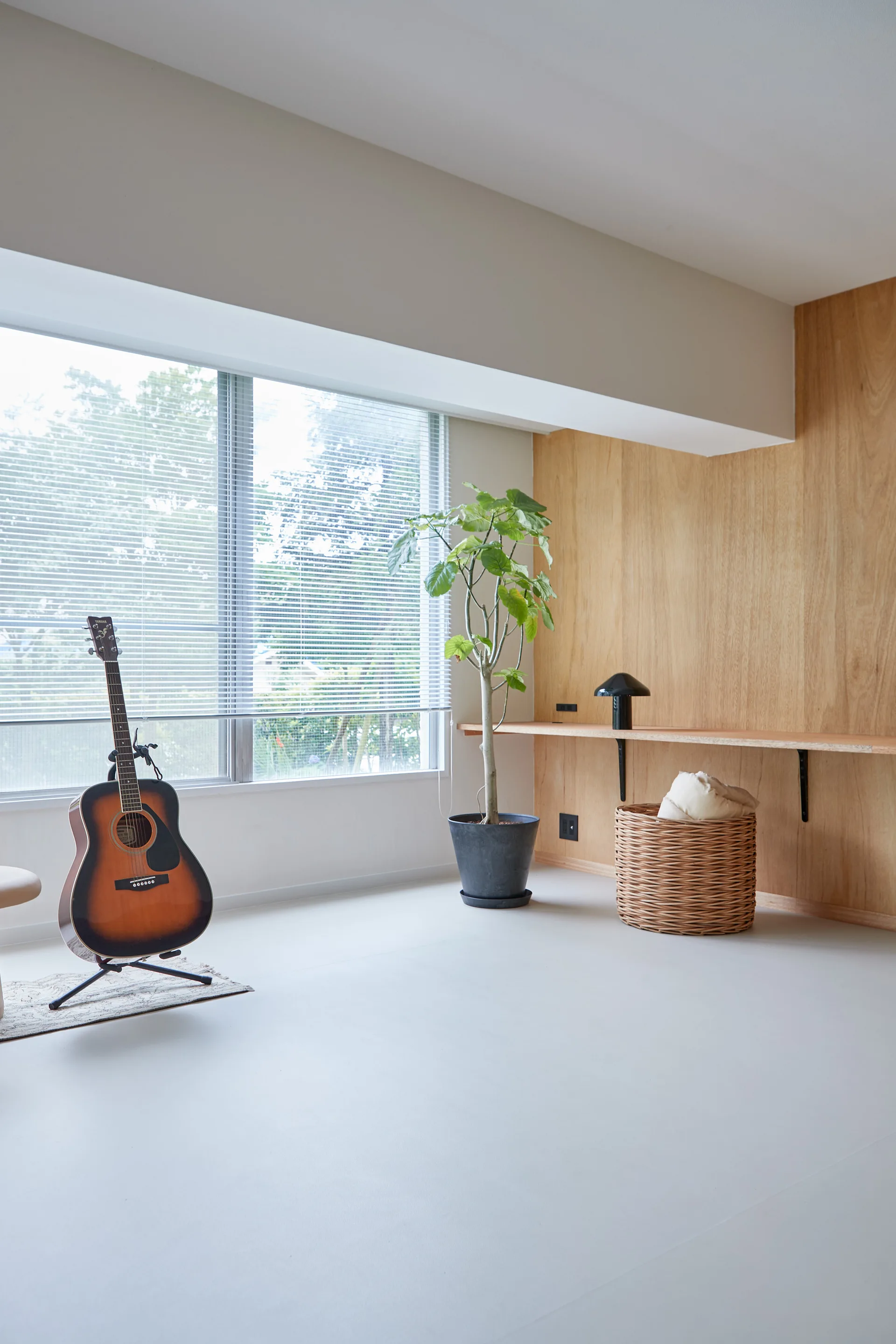 An acoustic guitar on a stand sits in a minimalist room featuring a large window with blinds, a wood-paneled wall, and a long floating shelf.