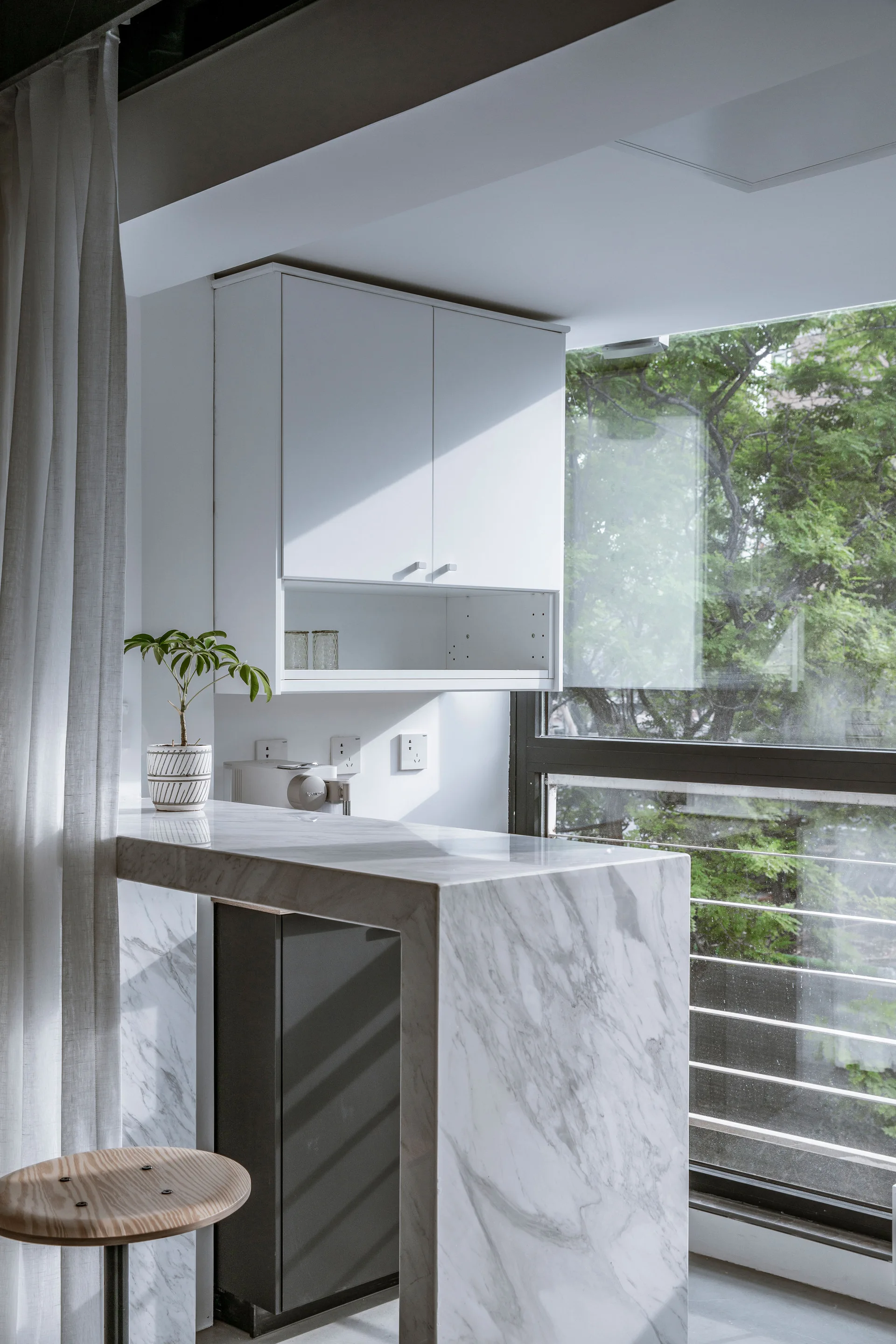 A bright kitchen features a white veined marble waterfall counter with a wooden stool next to a large window and white upper cabinets.