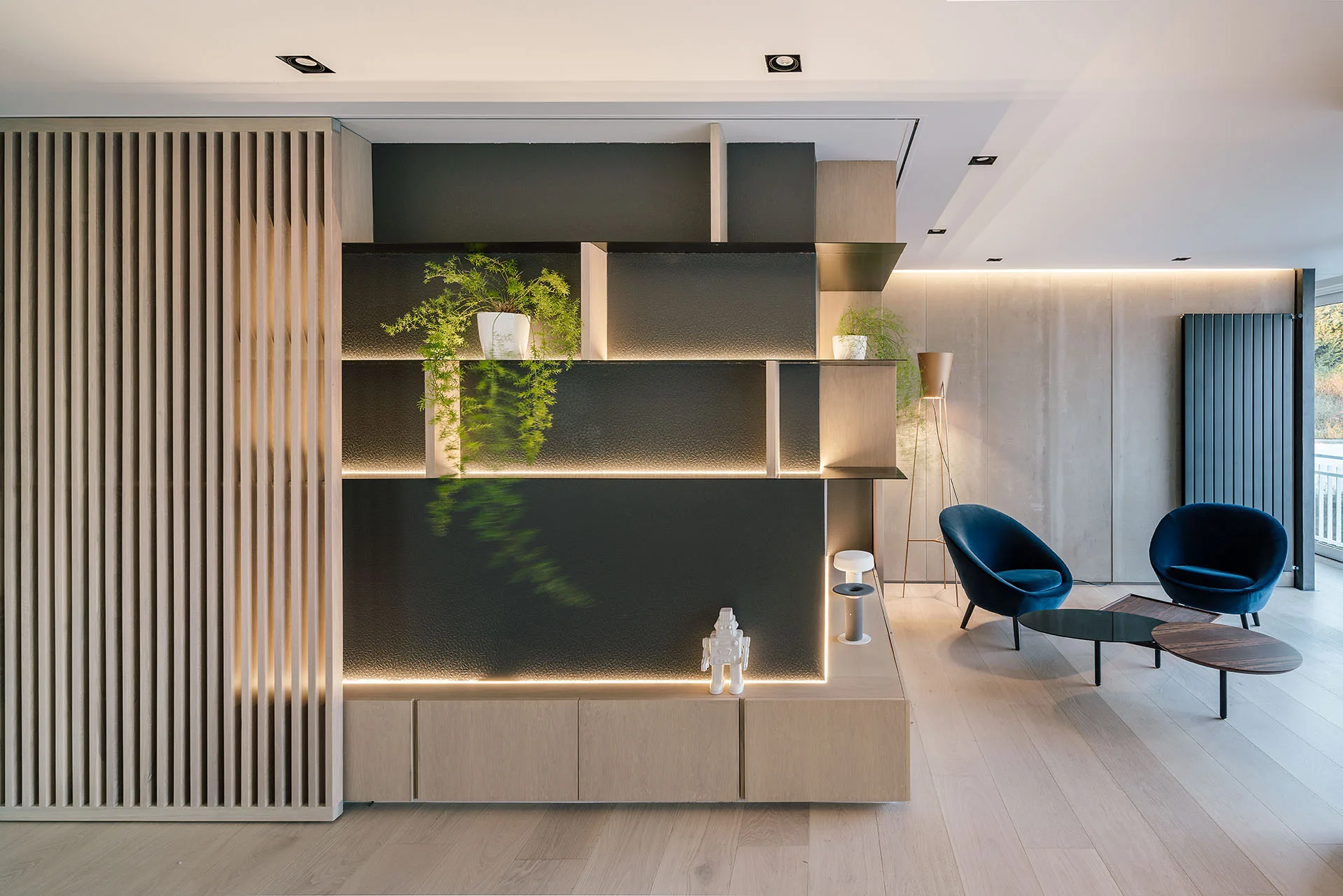 A contemporary lounge features a custom backlit wood shelving unit alongside a vertical slatted divider and two navy blue velvet armchairs.