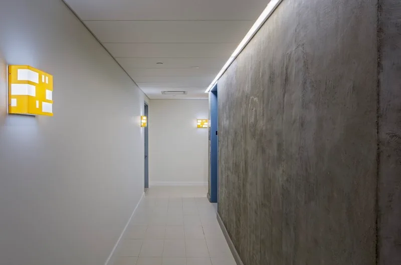 A minimalist hallway featuring a smooth white wall with yellow box sconces opposite a textured grey concrete wall under a paneled ceiling.