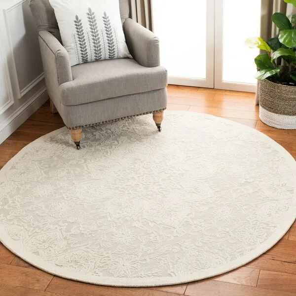 A round ivory damask patterned rug lies on a medium brown wood floor next to a grey upholstered armchair and a potted plant.