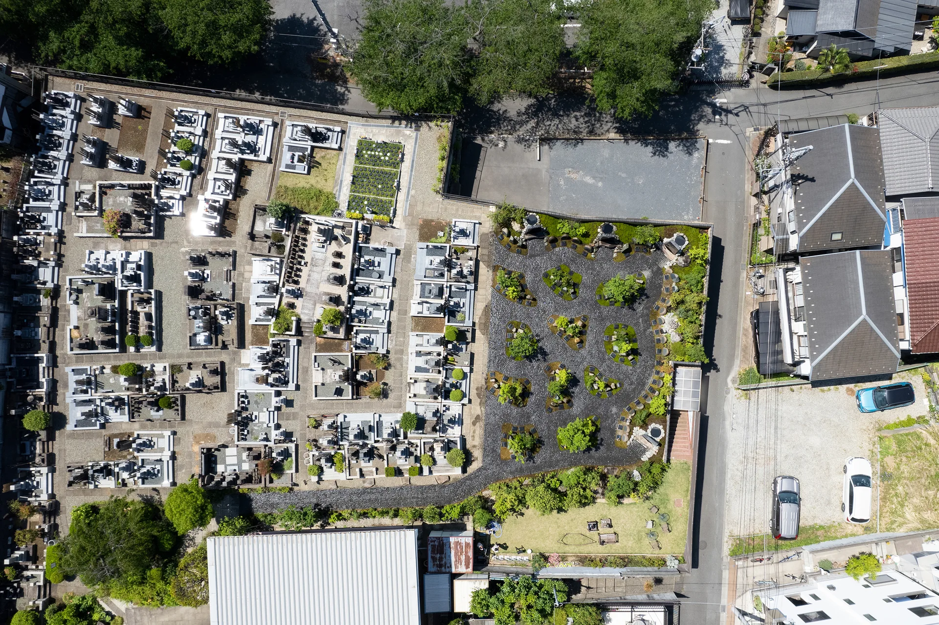 An aerial view of a structured landscape garden featuring dark gravel pathways and green tree clusters next to a traditional cemetery.
