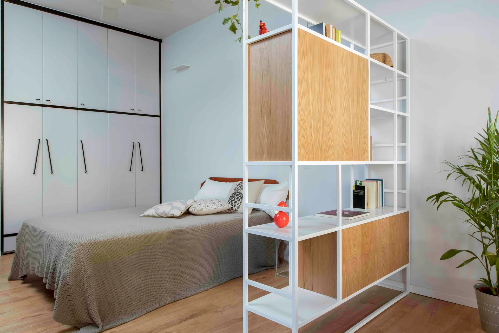 A modern bedroom features a grey bed next to a white metal frame room divider with wood cabinets and a built-in wardrobe in the background.