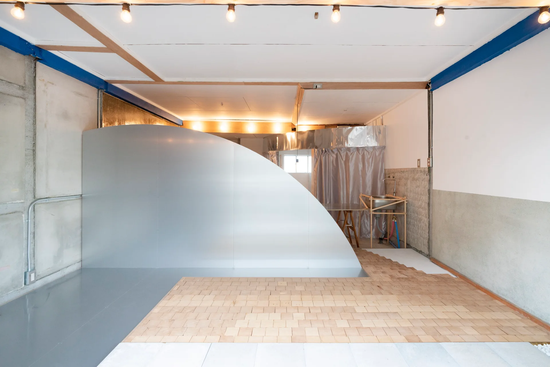 A raw industrial studio space featuring a large curved silver partition, mixed wood and grey flooring, and string lighting on the ceiling.