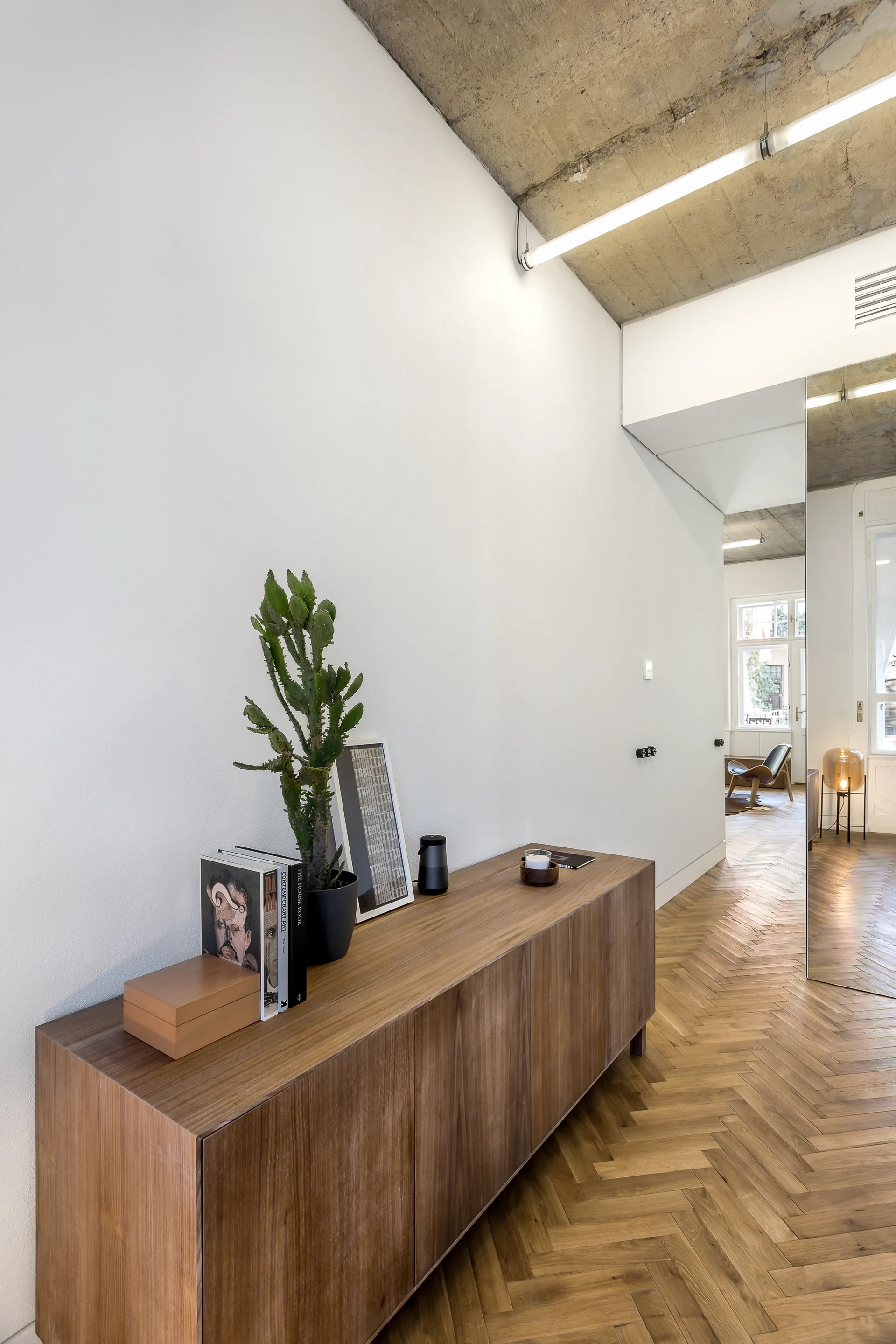 A long wooden sideboard stands on herringbone parquet flooring in a hallway with an exposed concrete ceiling and a floor-to-ceiling mirror.