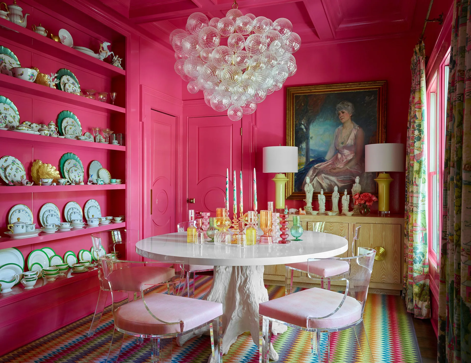 A dining room featuring high-gloss hot pink walls, a white round table, clear acrylic chairs with pink seats, and a large bubble glass chandelier.