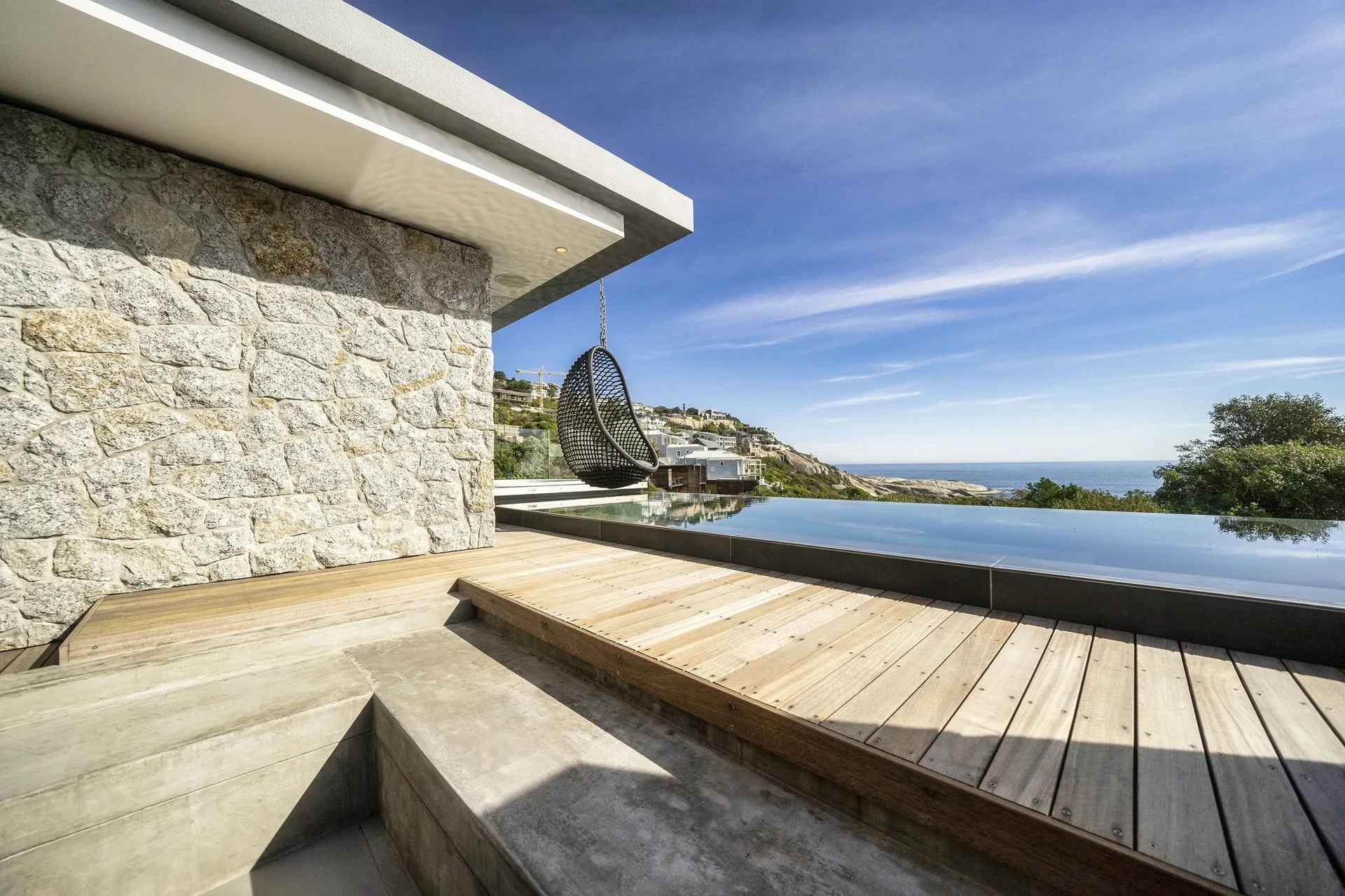 A modern wooden terrace features an infinity pool, a textured stone wall, and a black hanging chair overlooking a coastal hillside landscape.