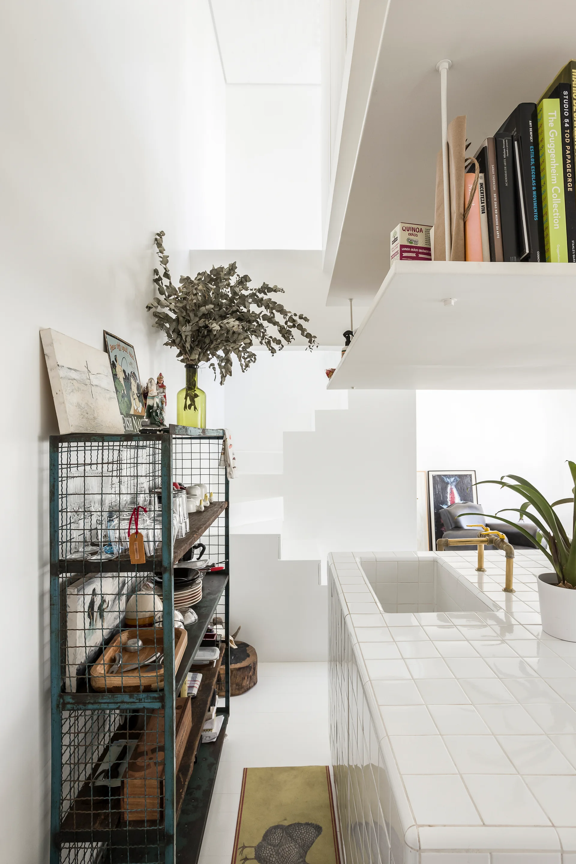 A white kitchen featuring a tiled island with a brass faucet, a distressed blue metal shelving unit, and a sculptural staircase in the background.
