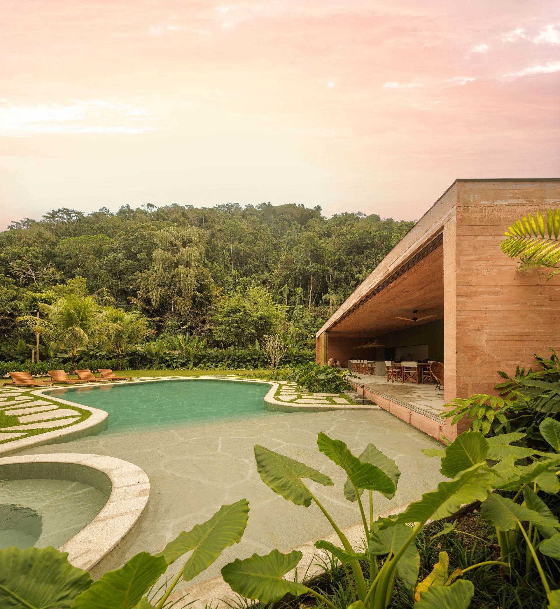 A curved outdoor swimming pool and flagstone patio next to a covered modern pavilion with terracotta walls under a warm sunset sky.