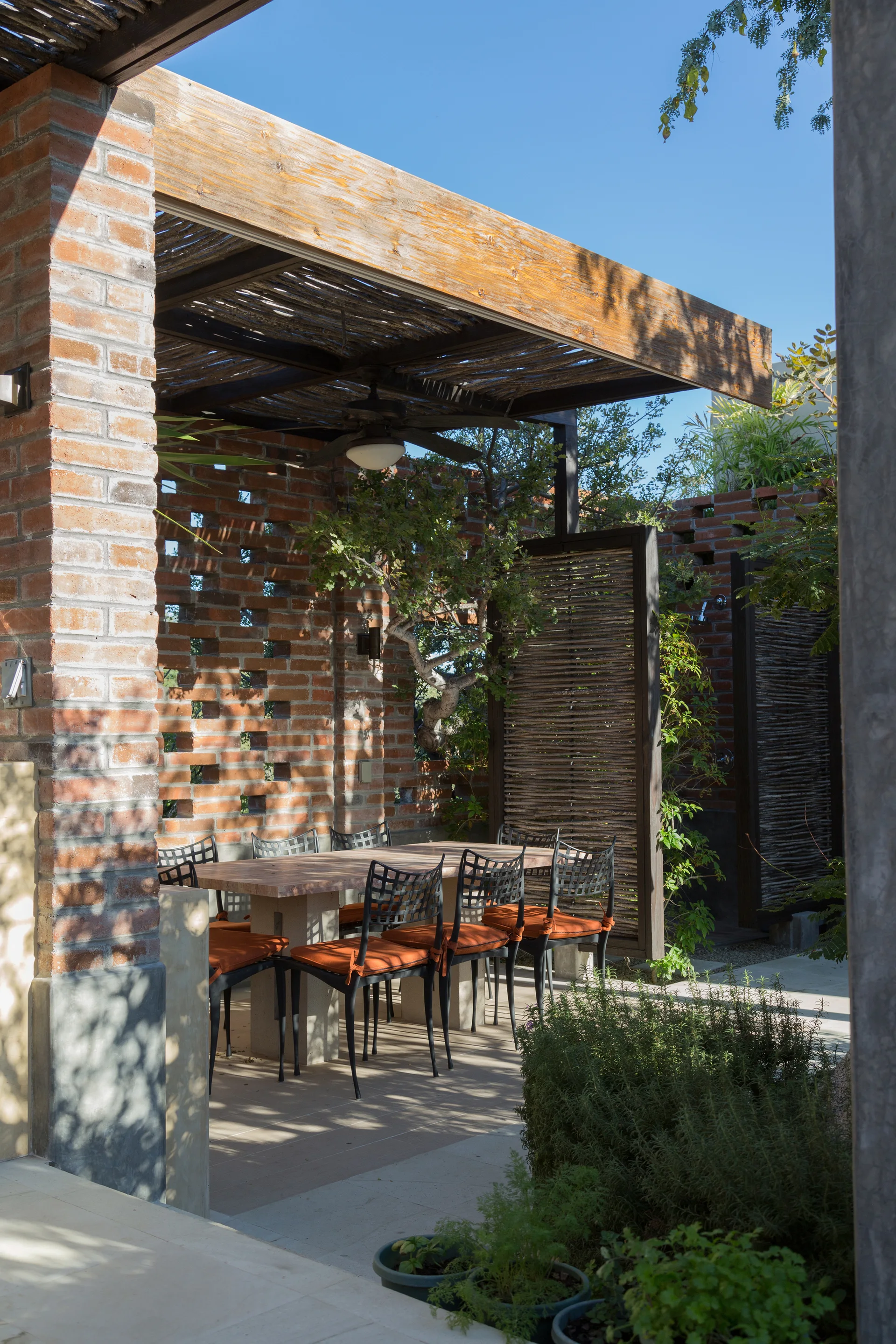 A rustic outdoor dining area features a stone table and black chairs with orange cushions beneath a wooden pergola beside a perforated brick screen wall.