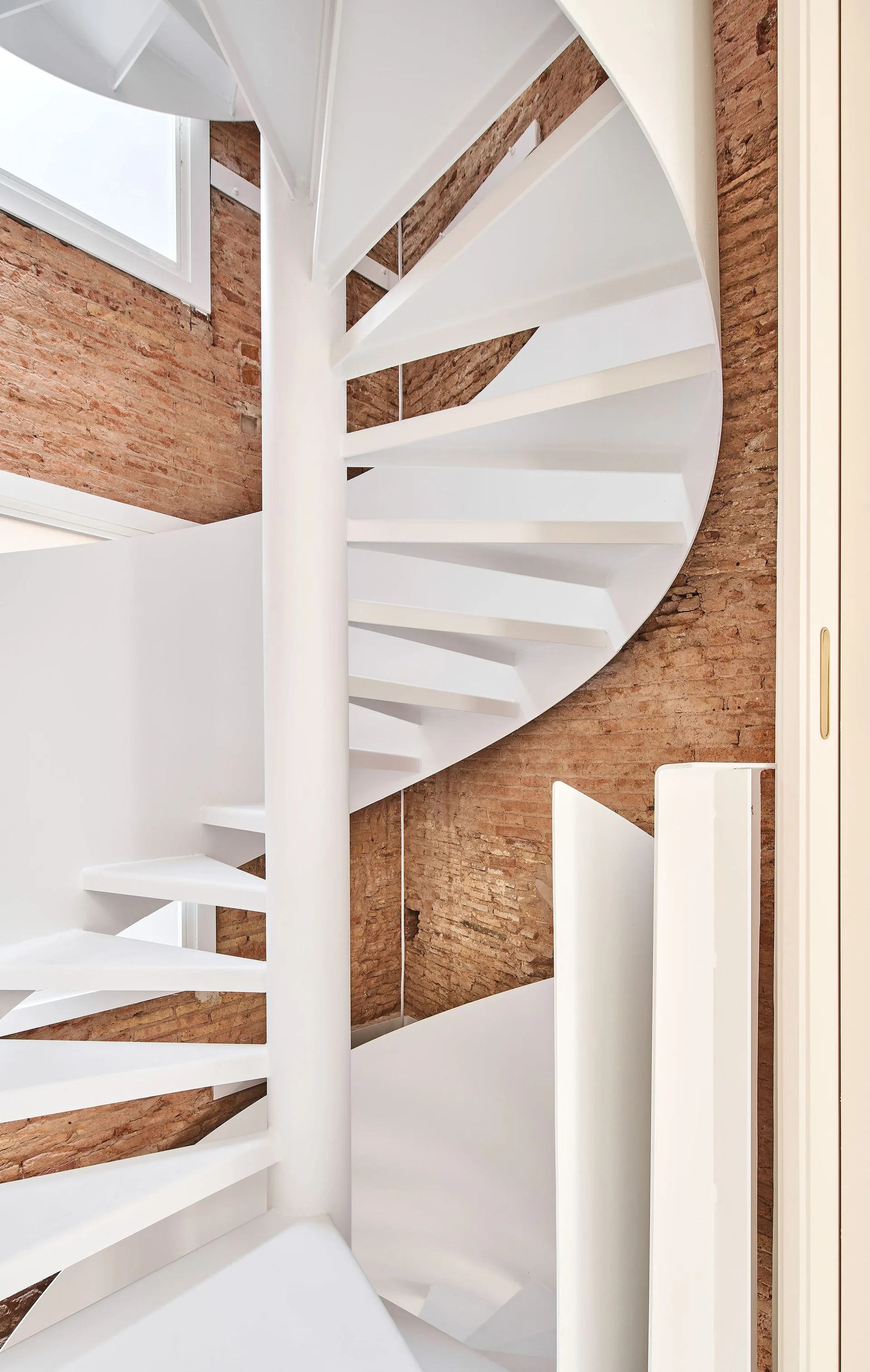 A minimalist white metal spiral staircase winds upward alongside a rustic exposed brick wall in a naturally lit room.