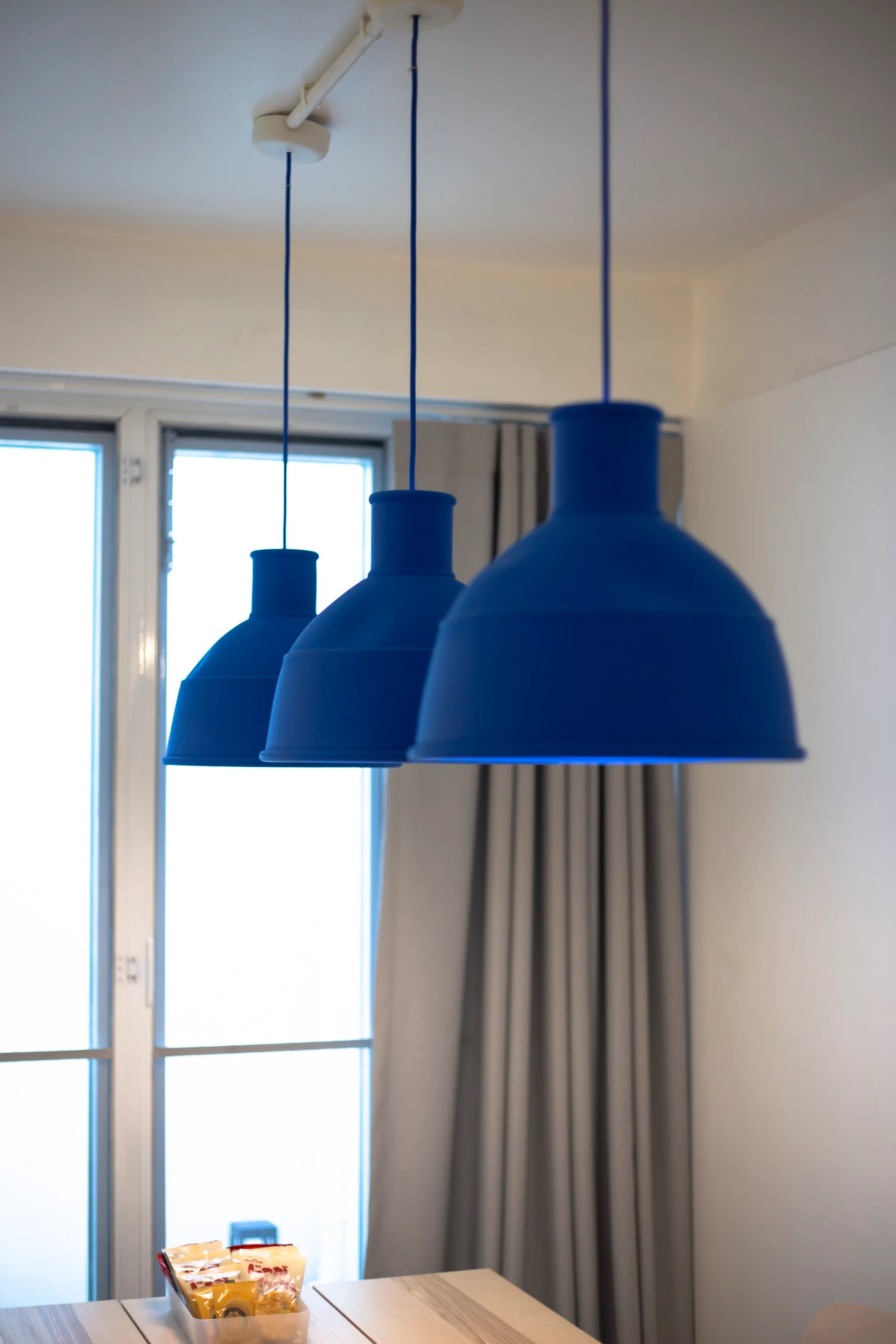 Three matte blue pendant lights hang in a row above a wooden dining table in front of a window with grey curtains.