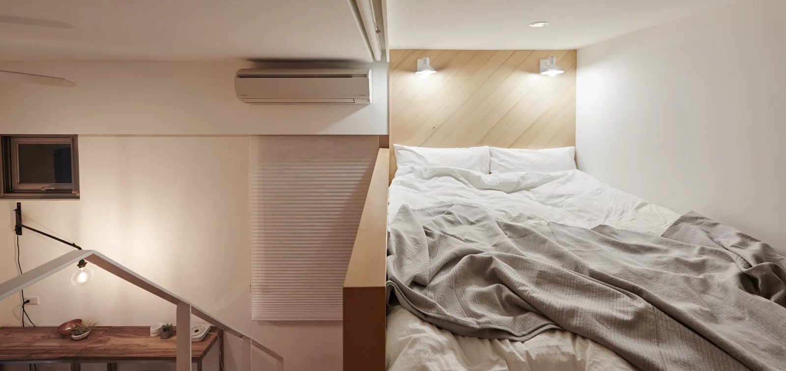 A compact loft bedroom features a bed with white bedding, two mounted sconces, and a diagonal light wood paneled headboard.
