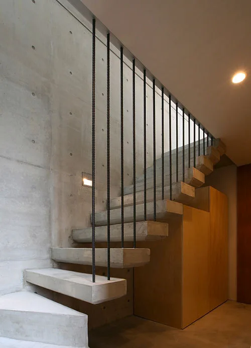 A minimalist floating concrete staircase with black vertical rod railings is set against an exposed concrete wall with wood under-stair cabinets.