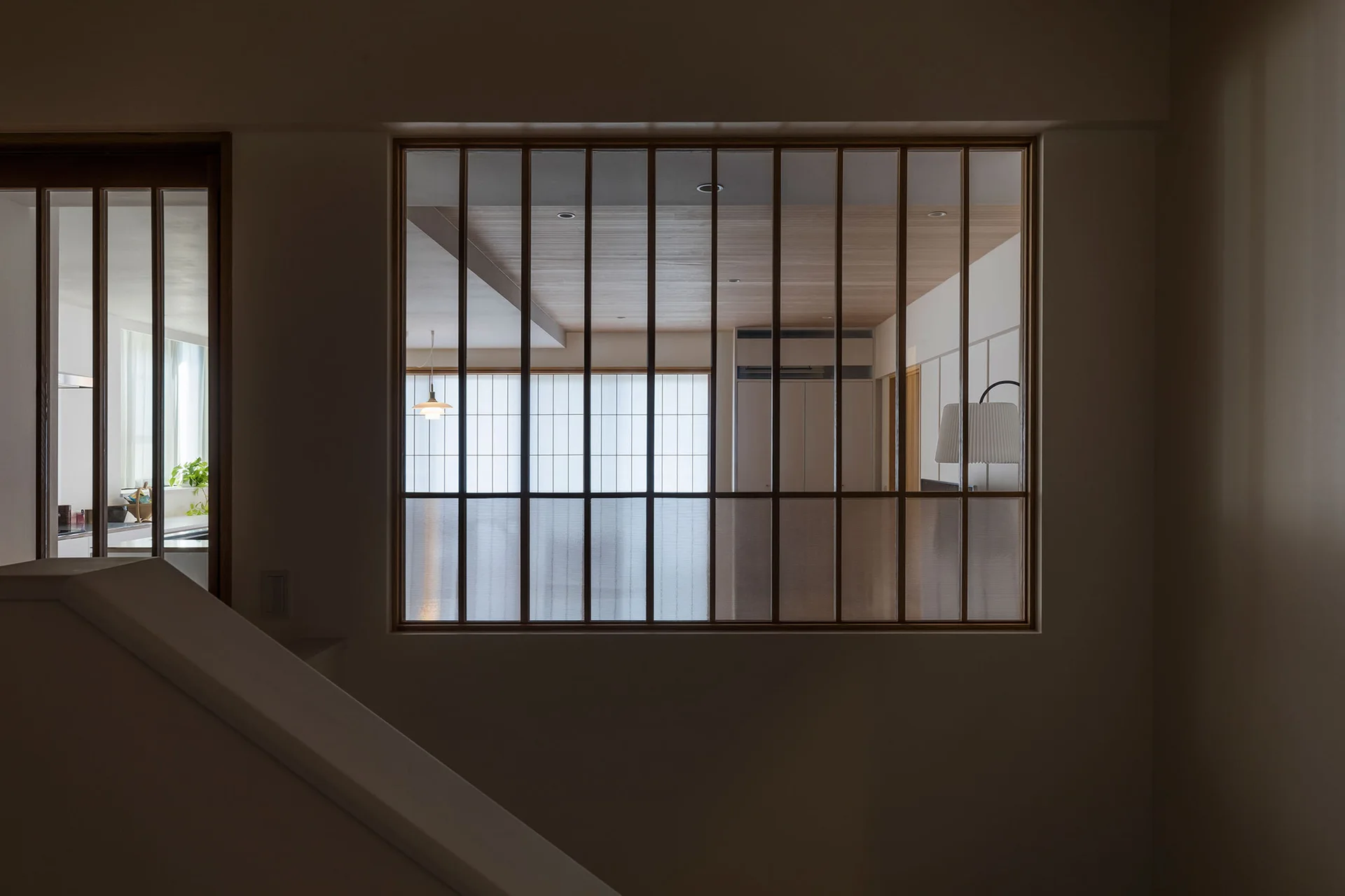 A dark hallway looking through a wood-framed gridded glass partition into a brightly lit living room with wood ceiling panels.
