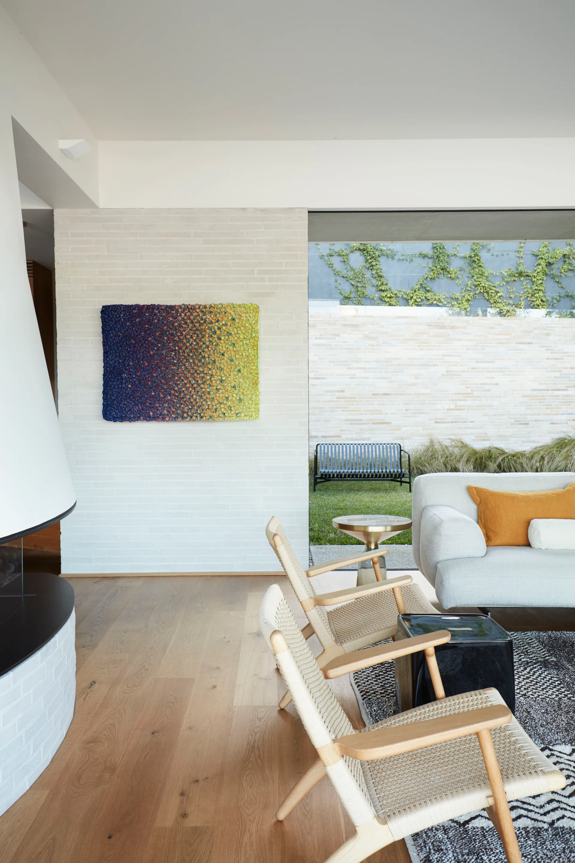 A bright living room features woven-seat lounge chairs, a grey sofa, and a colorful textured art piece mounted on a white brick wall.