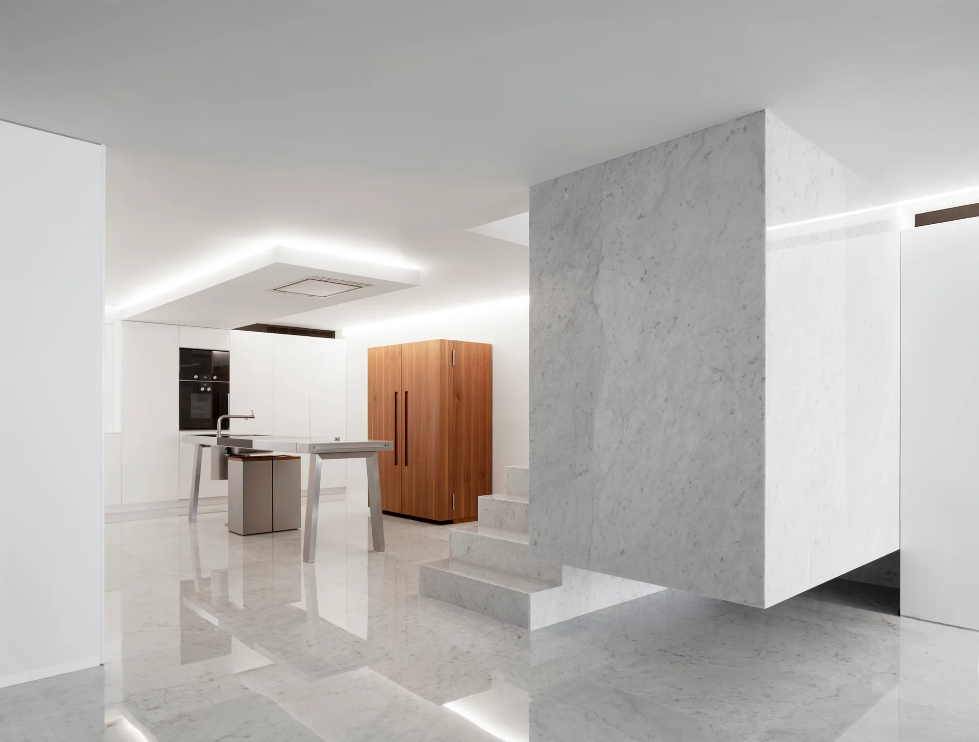 A minimalist white kitchen featuring a large marble monolith staircase, a metal island, and a tall wood cabinet on polished stone floors.
