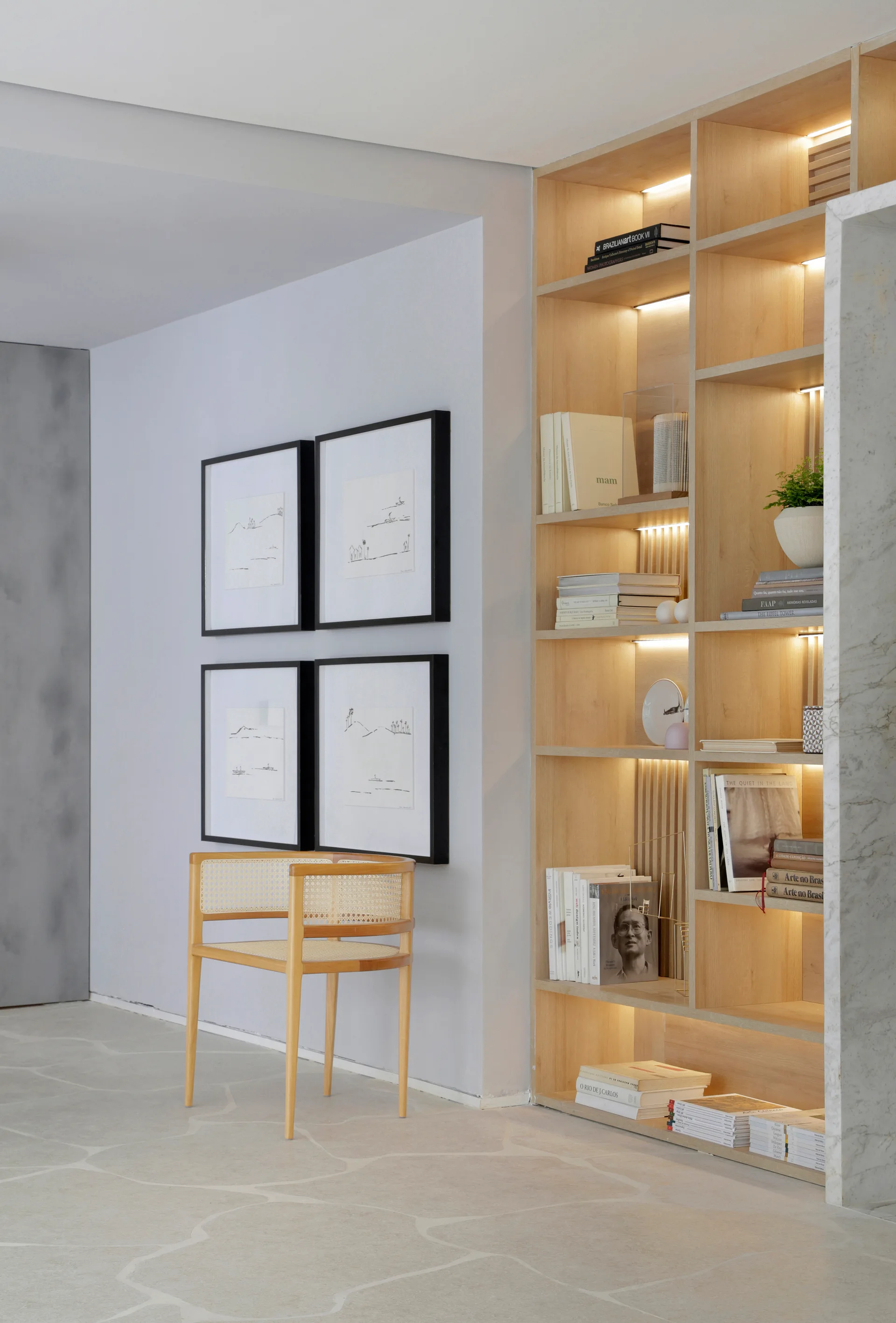 A light wood built-in bookcase with integrated lighting stands next to a gray wall decorated with four black-framed art pieces and a cane chair.