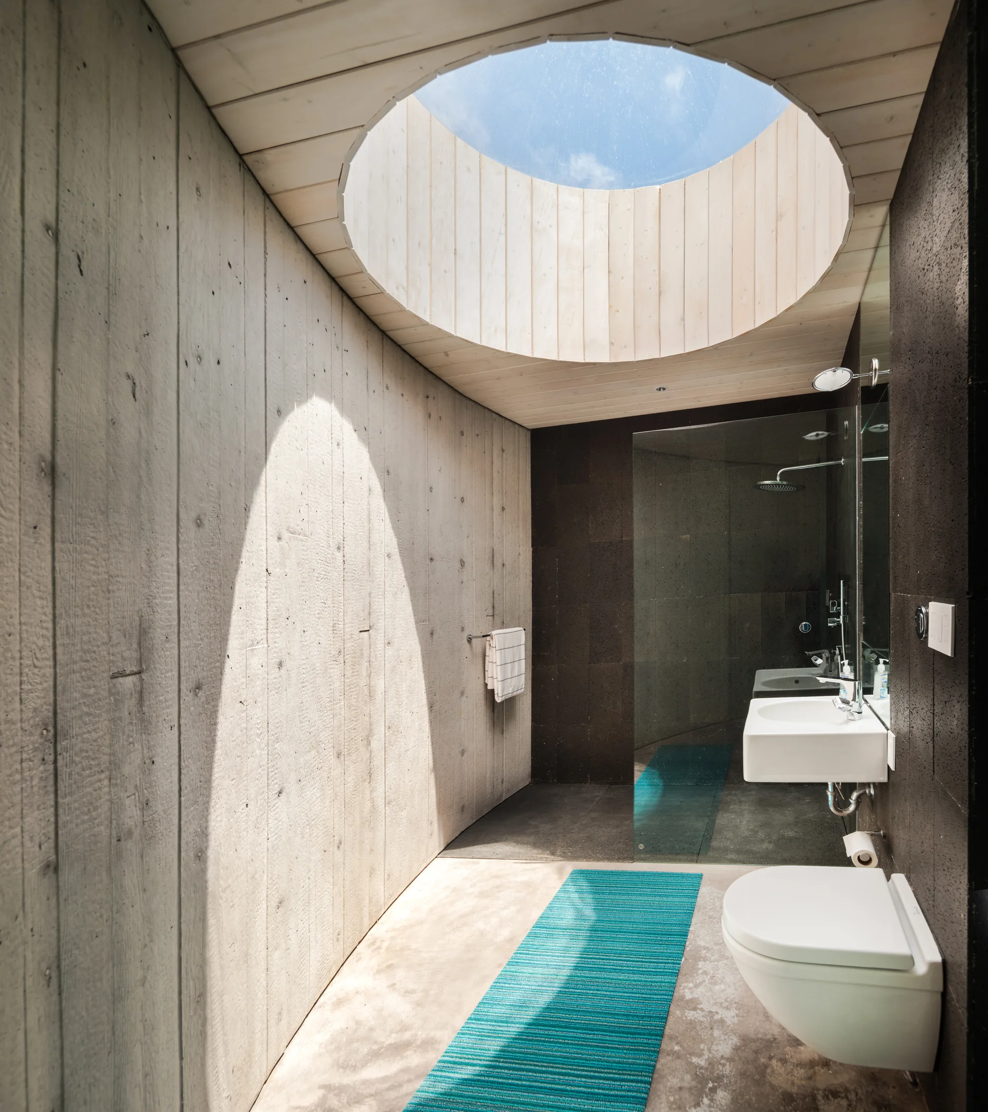 A modern bathroom featuring board-marked concrete walls, a large circular skylight overhead, and a bright teal runner rug on the floor.
