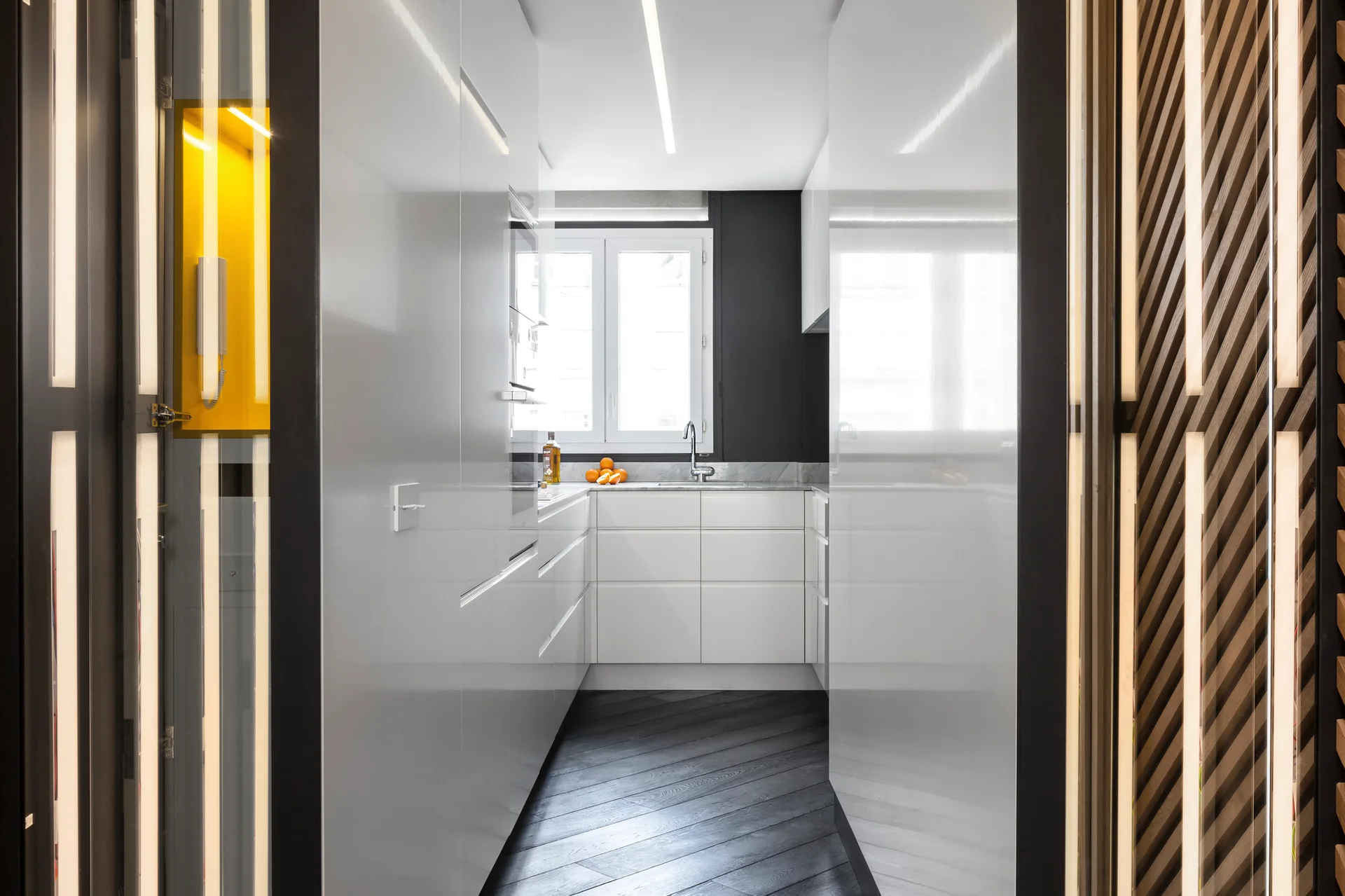 A narrow kitchen features high-gloss white cabinetry, a central window, dark herringbone wood floors, and a slatted timber wall partition.