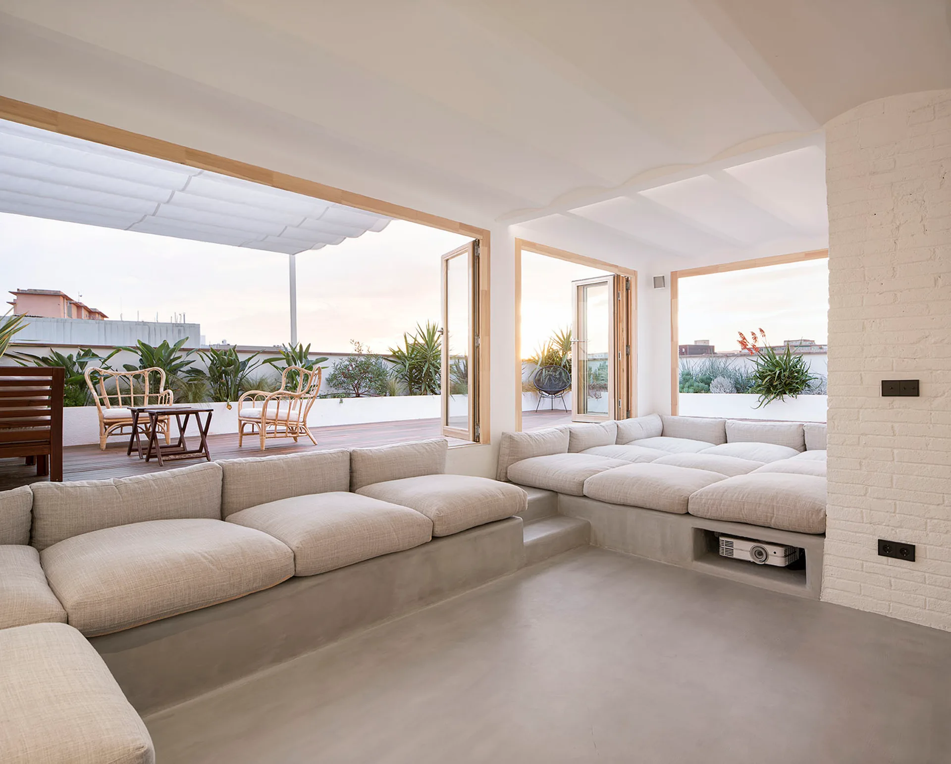 A minimalist living room features low built-in concrete seating with beige cushions next to folding glass doors that open to a sunny terrace.