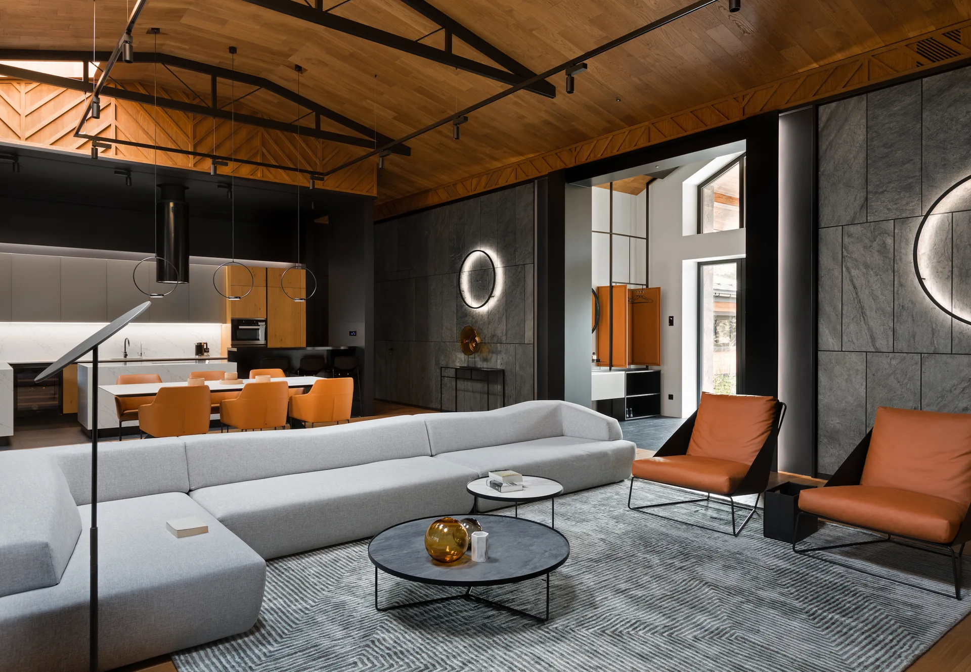 A modern open-plan living area featuring a light gray sectional sofa, orange leather chairs, and circular wall sconces on stone panels.