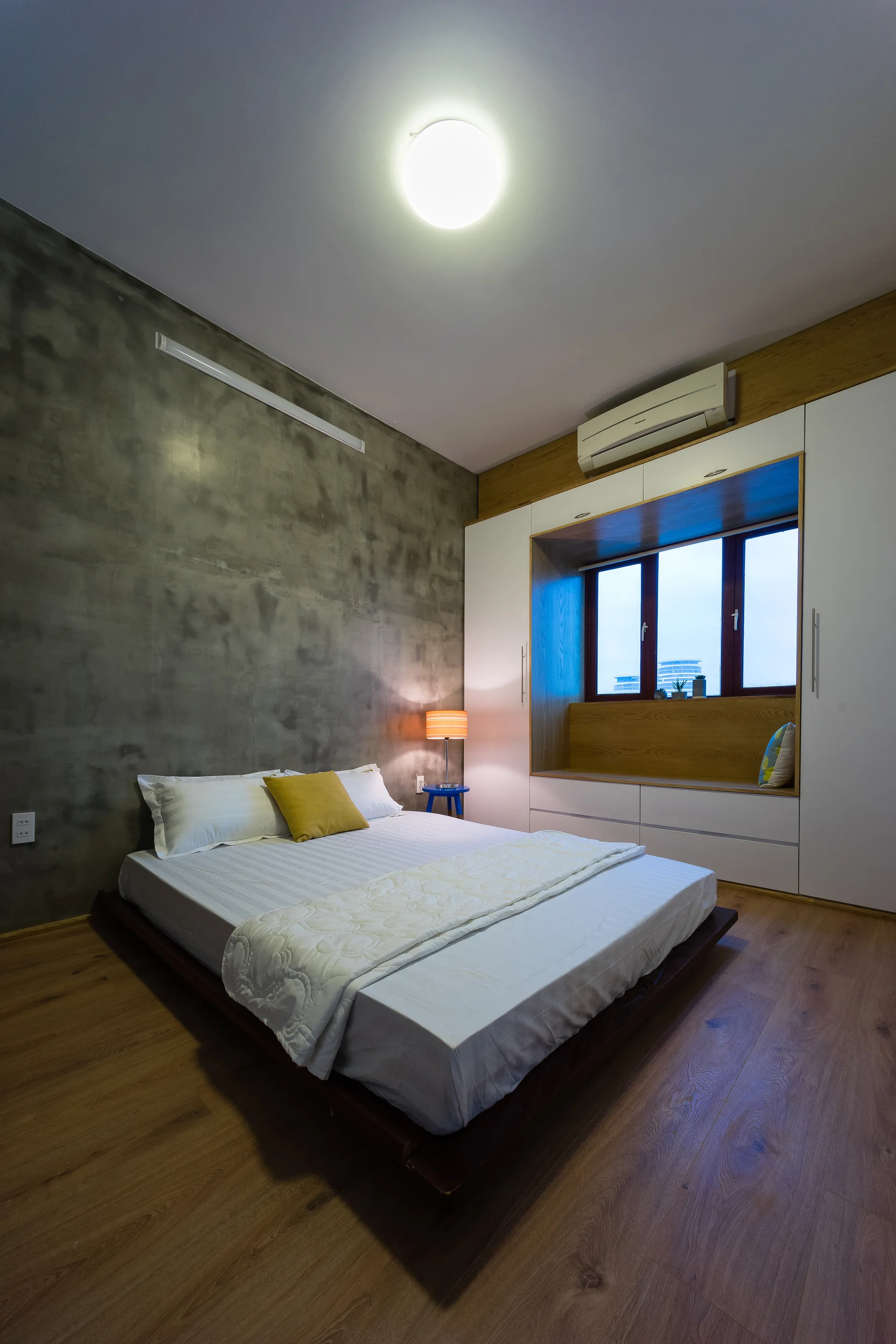 A modern bedroom features a low platform bed against a grey concrete wall and a built-in white wardrobe system framing a wooden window seat.