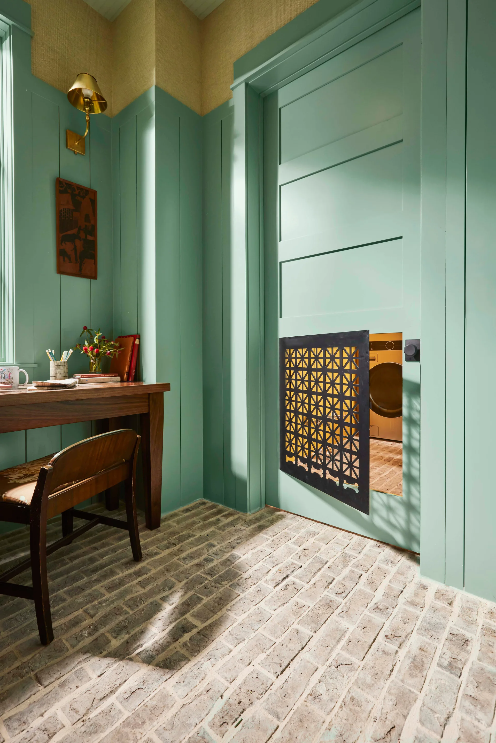 A sage green mudroom featuring a textured brick floor, a wooden writing desk, and a door with an integrated black geometric pet gate.