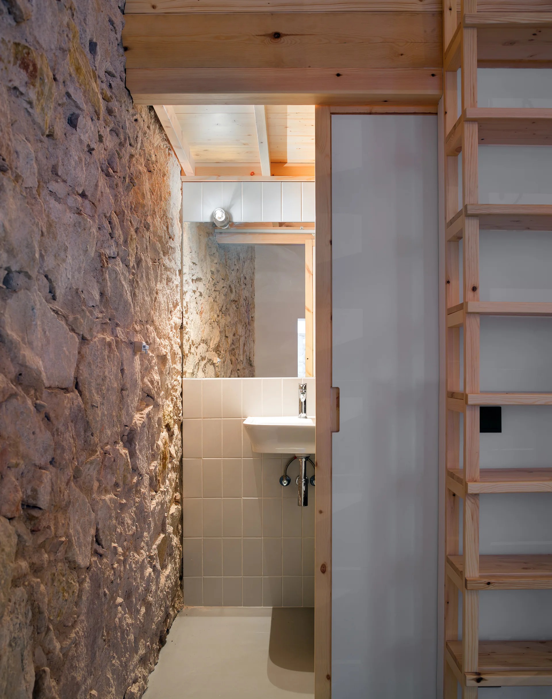 A compact bathroom features a wall-mounted sink with square white tiles, framed by a rugged exposed stone wall and natural wood ceiling beams.