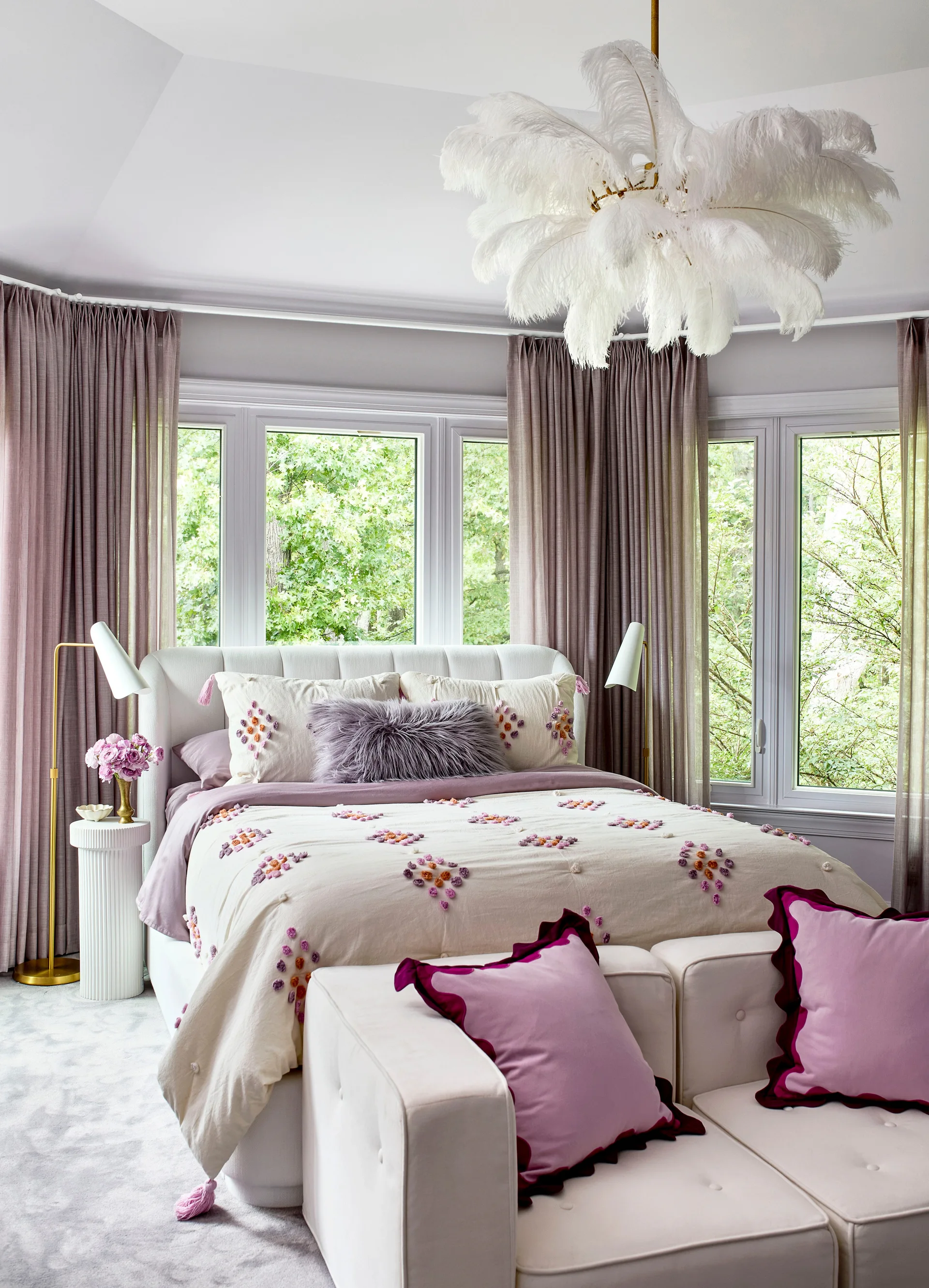 A bedroom features an upholstered bed with purple accent pillows, framed by mauve curtains, a white modular ottoman, and a fluffy feather chandelier.