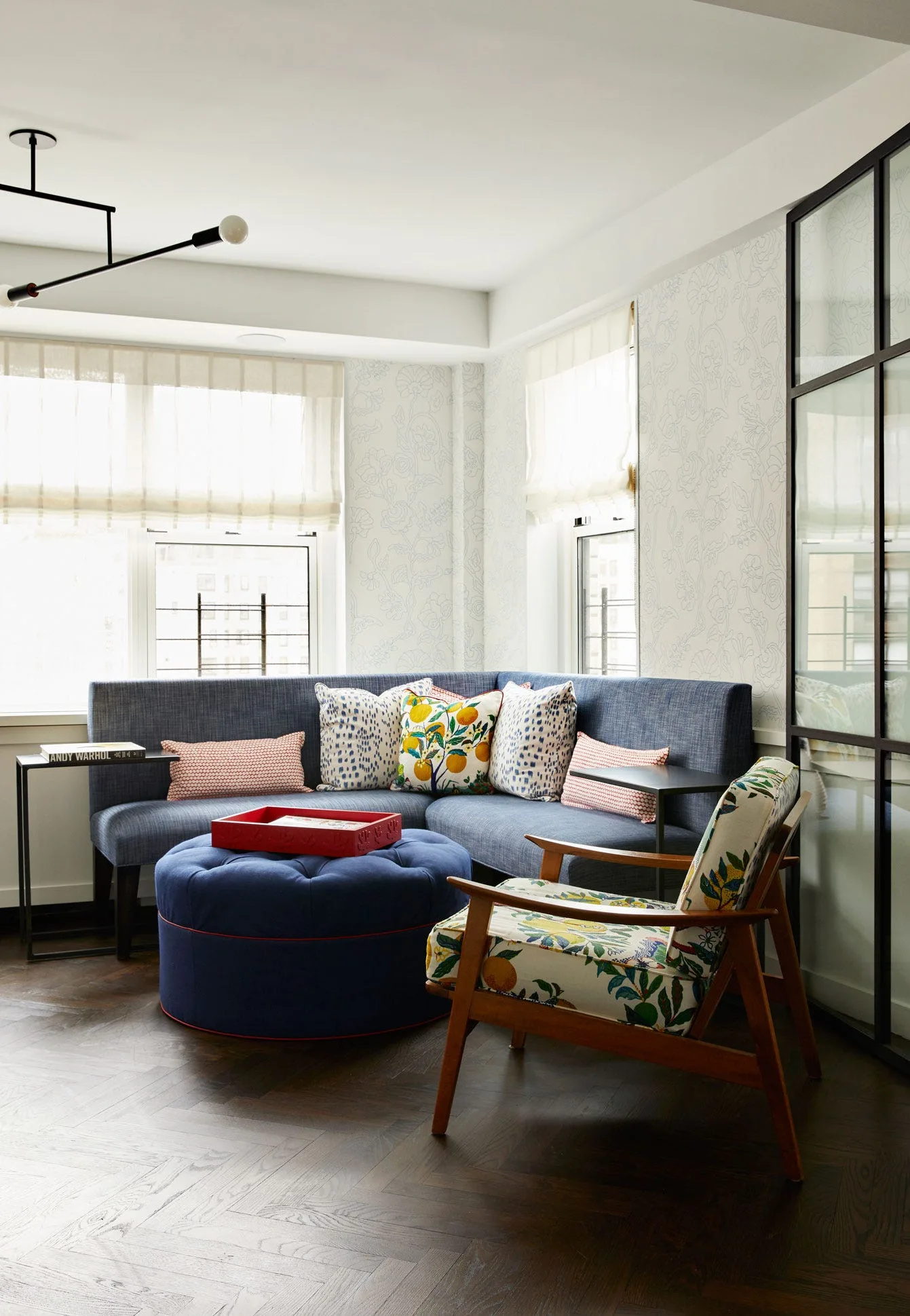 A cozy corner seating area with a blue sectional sofa, a round blue ottoman, a floral armchair, and light patterned wallpaper over herringbone floors.