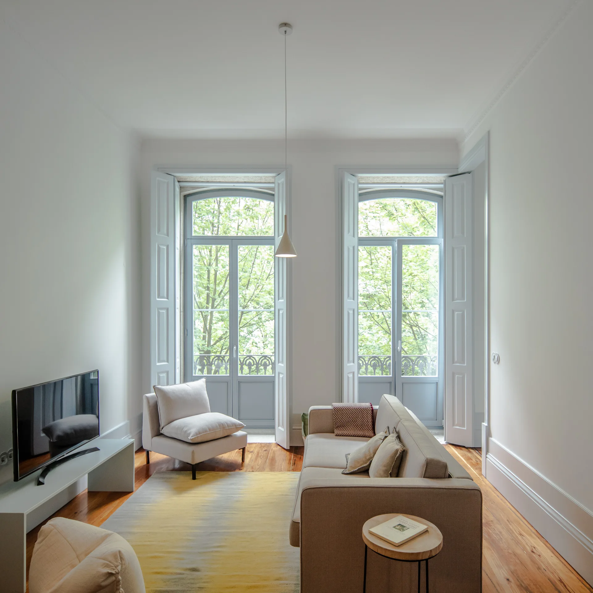 A bright living room features a beige sofa, a yellow-and-grey gradient rug, and tall French doors with soft blue-grey interior shutters.