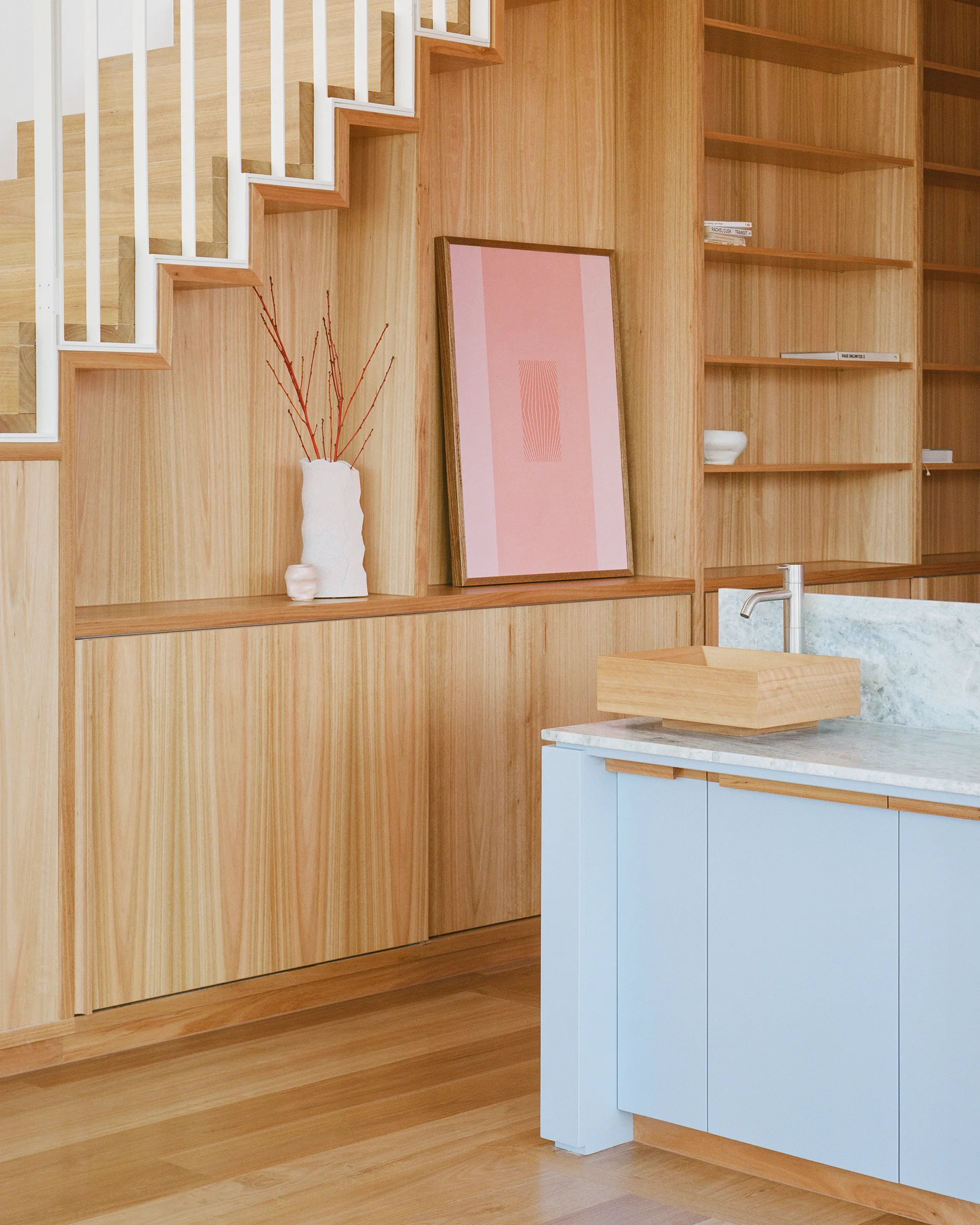A modern kitchen featuring extensive light wood wall paneling integrated with a staircase next to a light blue island with a marble countertop.