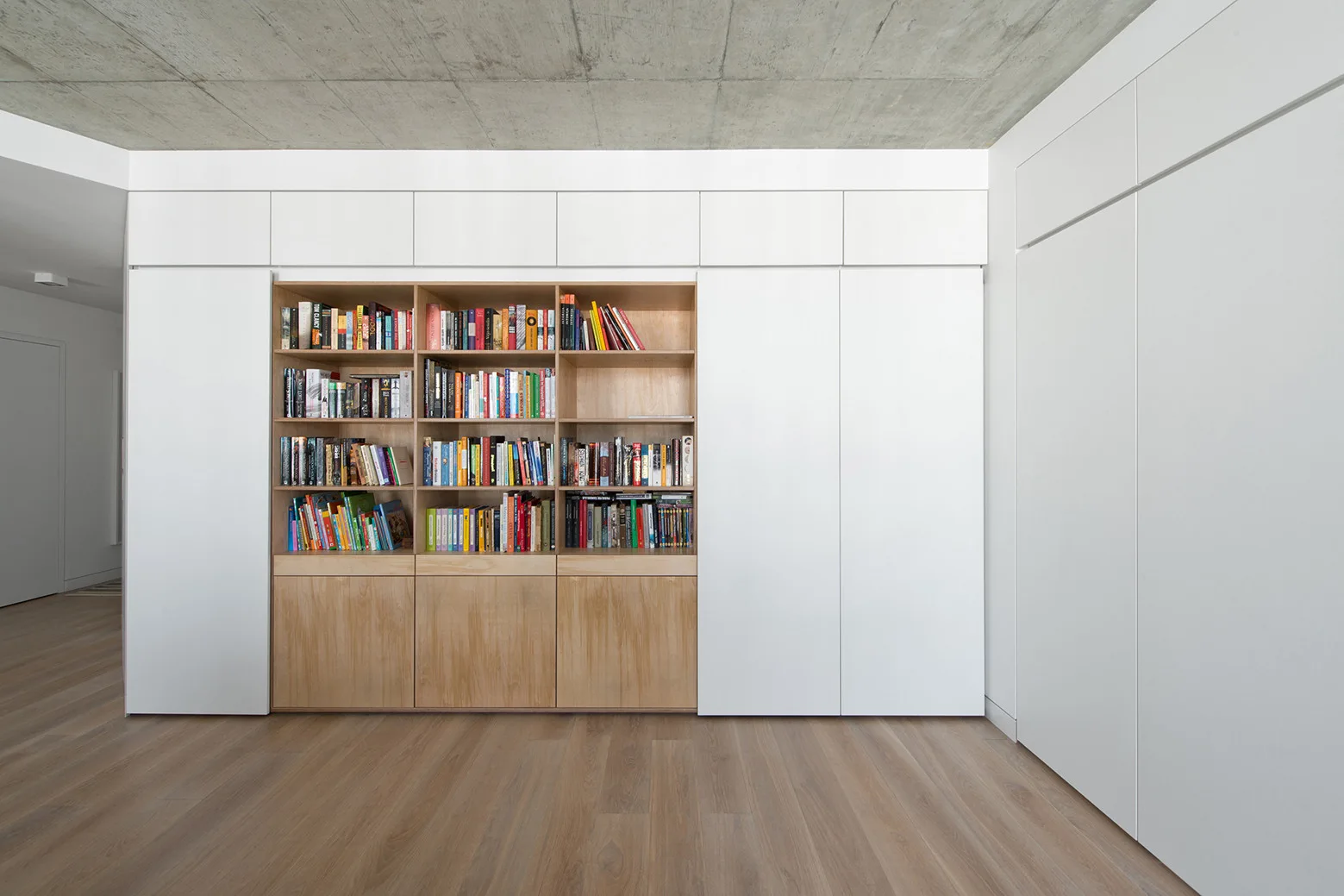 A floor-to-ceiling built-in wooden bookshelf filled with books is flanked by large white flat-panel storage doors beneath a raw concrete ceiling.