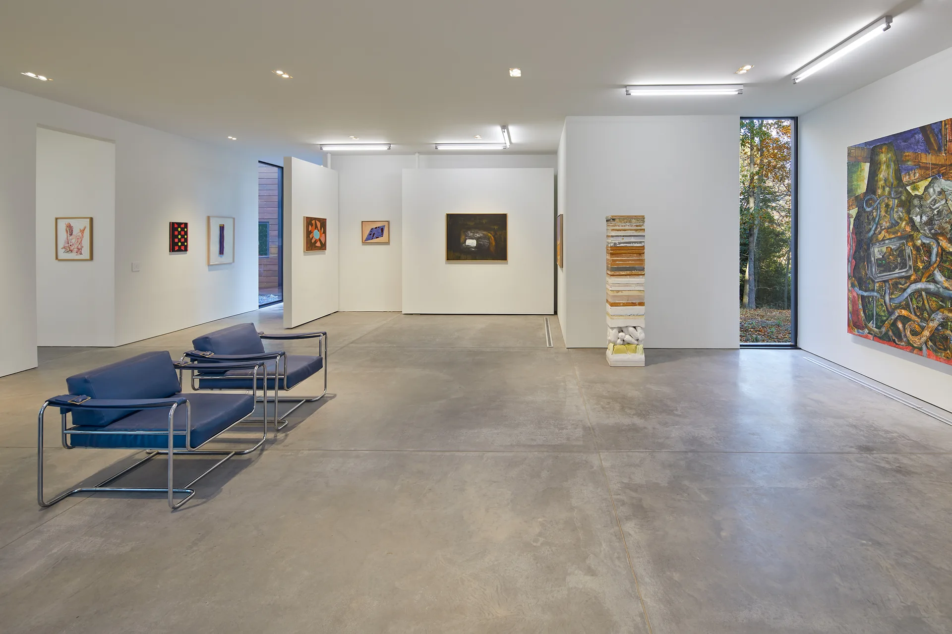 Two blue leather armchairs with metal frames sit on a polished concrete floor in a spacious white-walled art gallery displaying paintings.