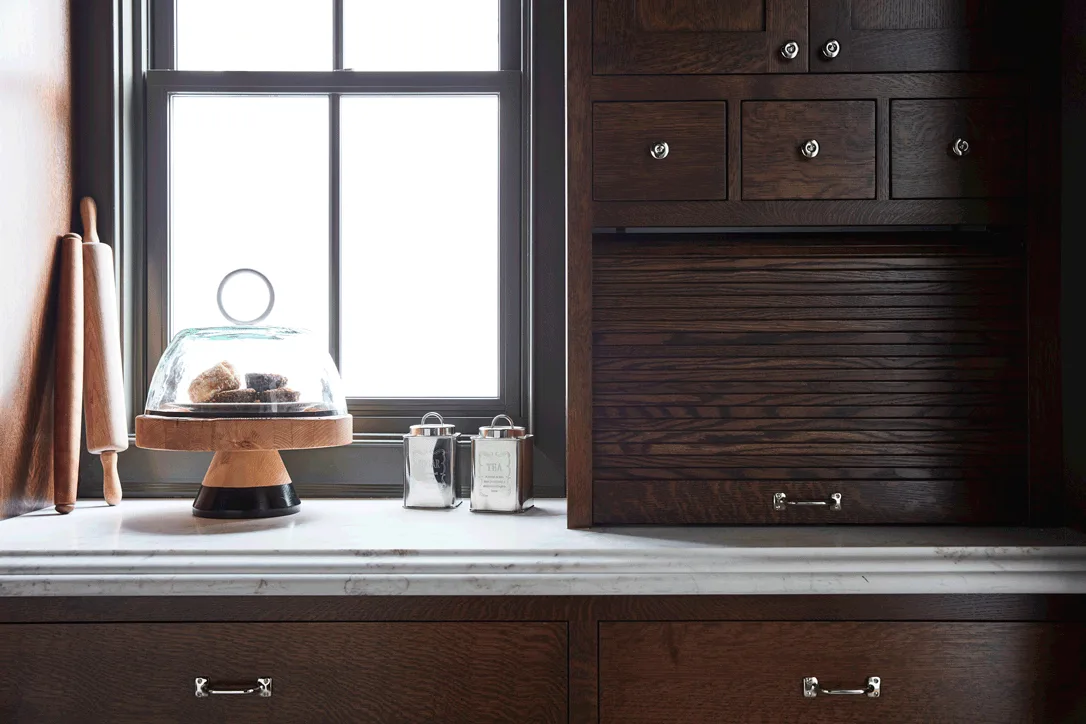 A close-up of dark wood kitchen cabinetry featuring a roll-up tambour door next to a window on a white veined marble countertop.