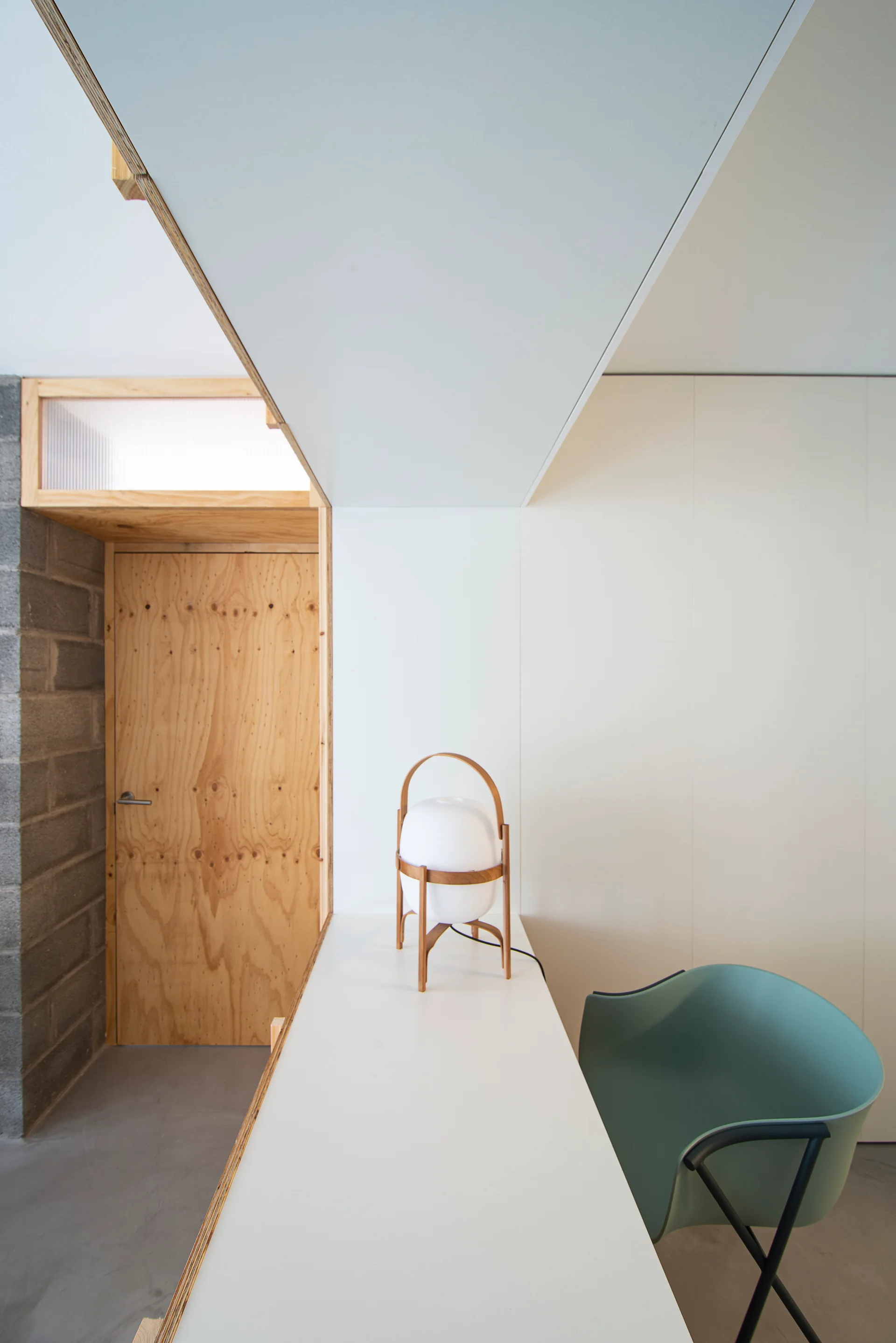A minimalist home office featuring a white built-in desk with a sage green chair, a lantern lamp, a plywood door, and a concrete block wall.