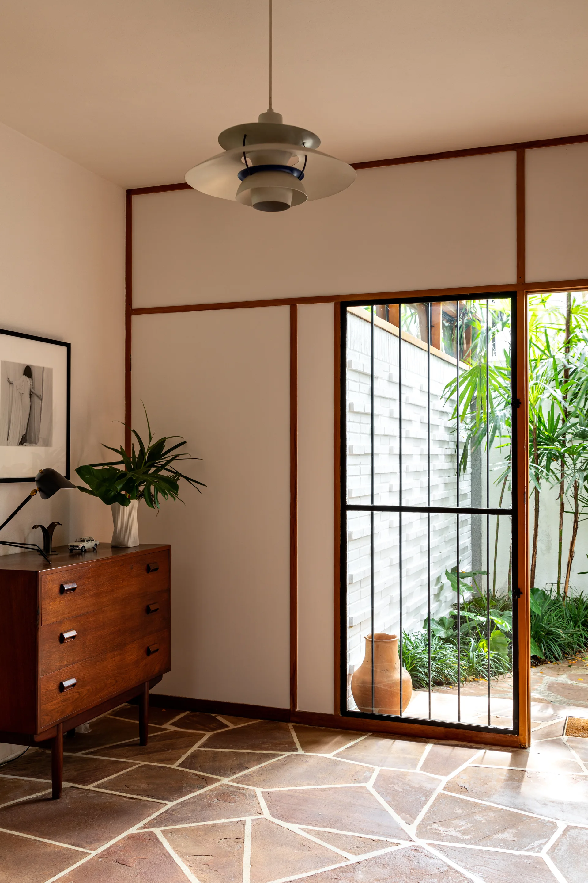 A mid-century modern interior featuring a wooden dresser, flagstone flooring, and a glass door opening to a green courtyard.