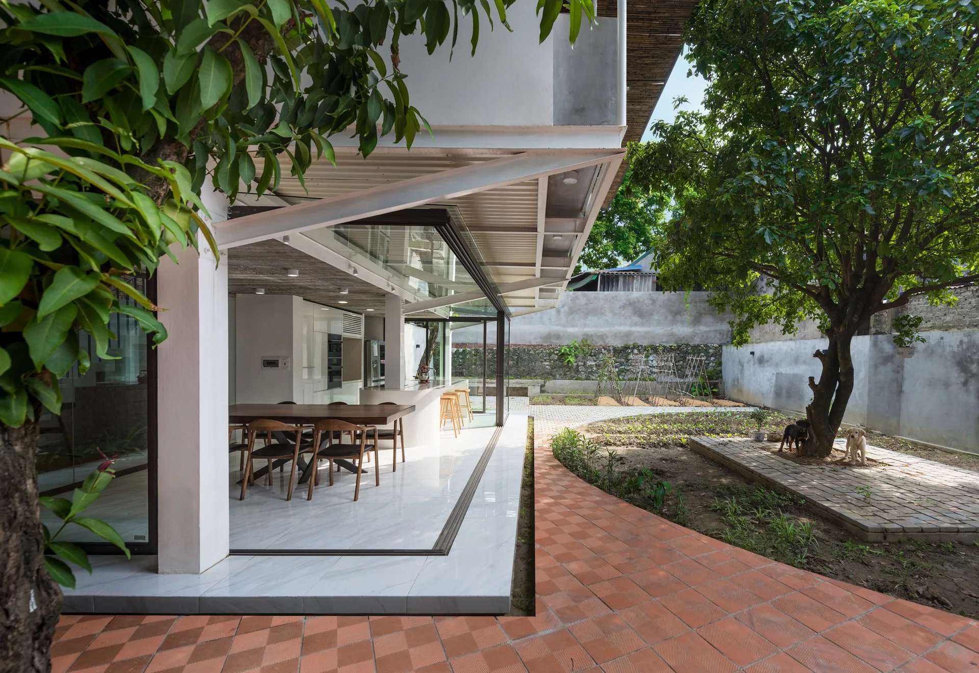 A modern kitchen and dining area with white floors and a dark wood table opens fully to a terracotta tiled patio and green garden.