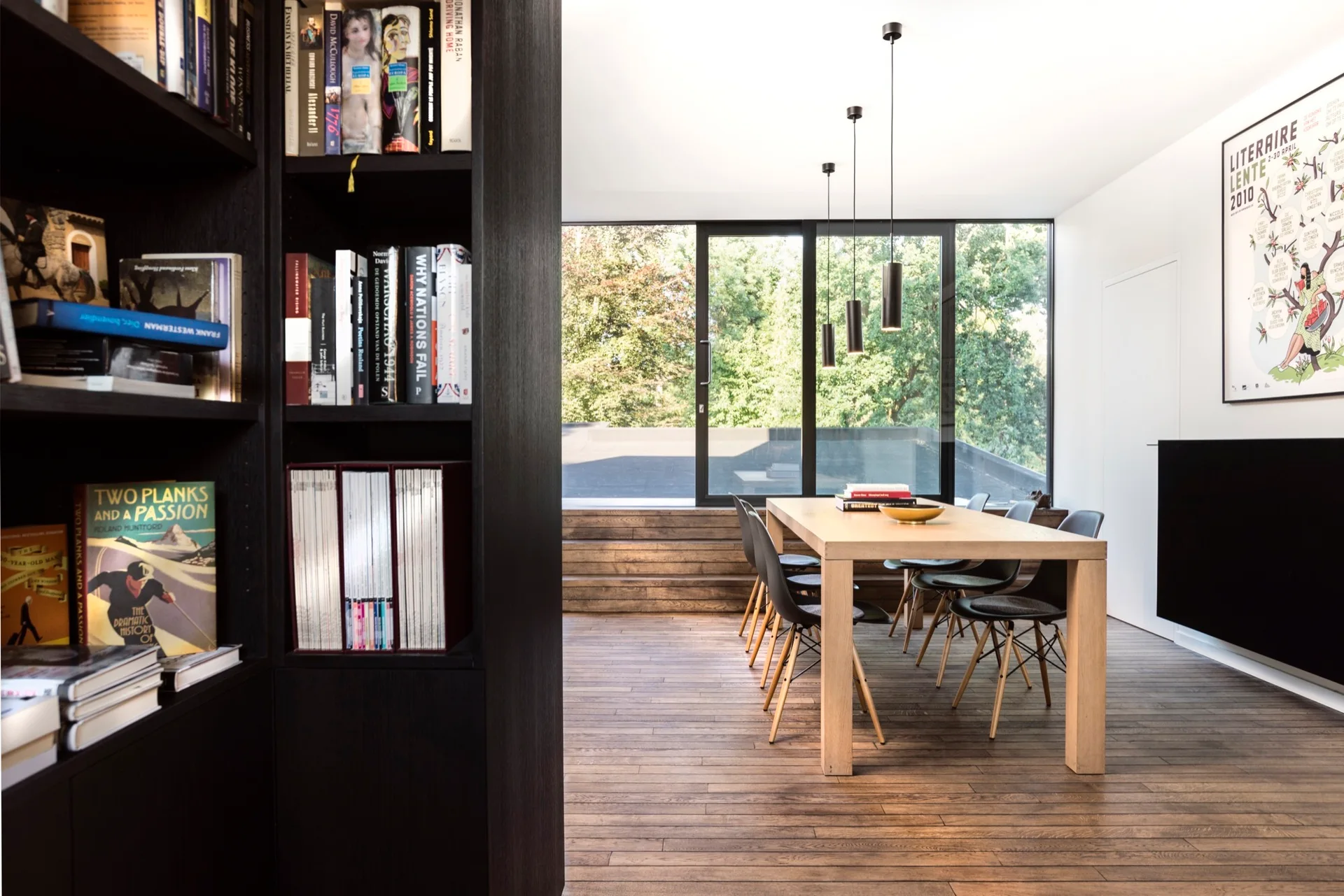 A minimalist dining room features a light wood table, black chairs, and a tall black bookcase in the foreground facing a glass window wall.