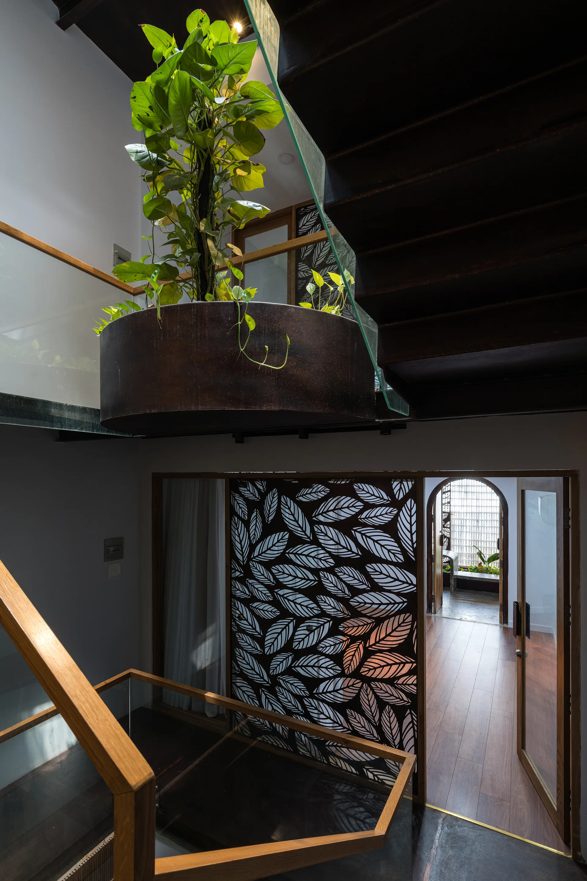 A modern stairwell features a suspended dark metal planter with green plants, a wood-trimmed glass railing, and a decorative leaf-patterned screen.