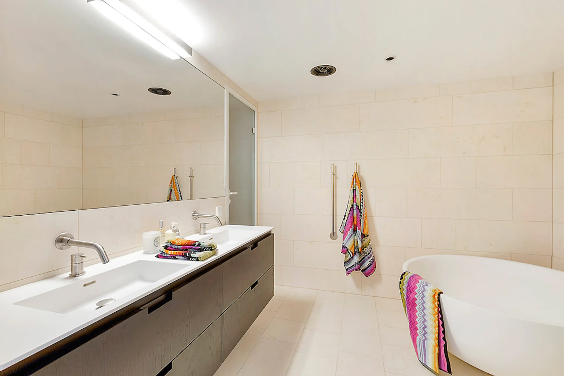 A modern bathroom featuring large format beige wall tiles, a dark double vanity with wall-mounted faucets, and a white freestanding tub.