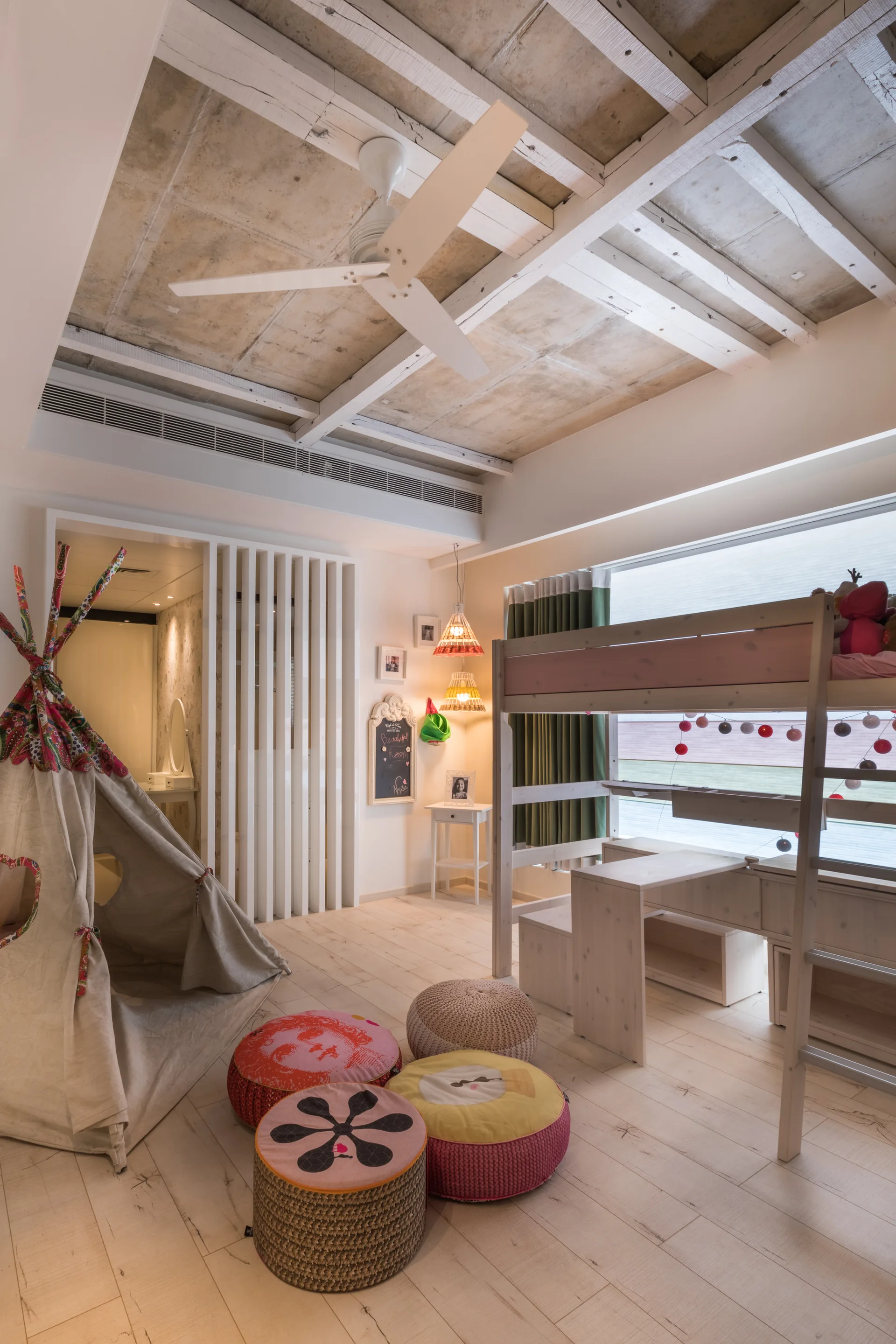 A children's bedroom features a light wood bunk bed with an integrated desk, a play teepee, colorful floor cushions, and a vertical slatted divider.