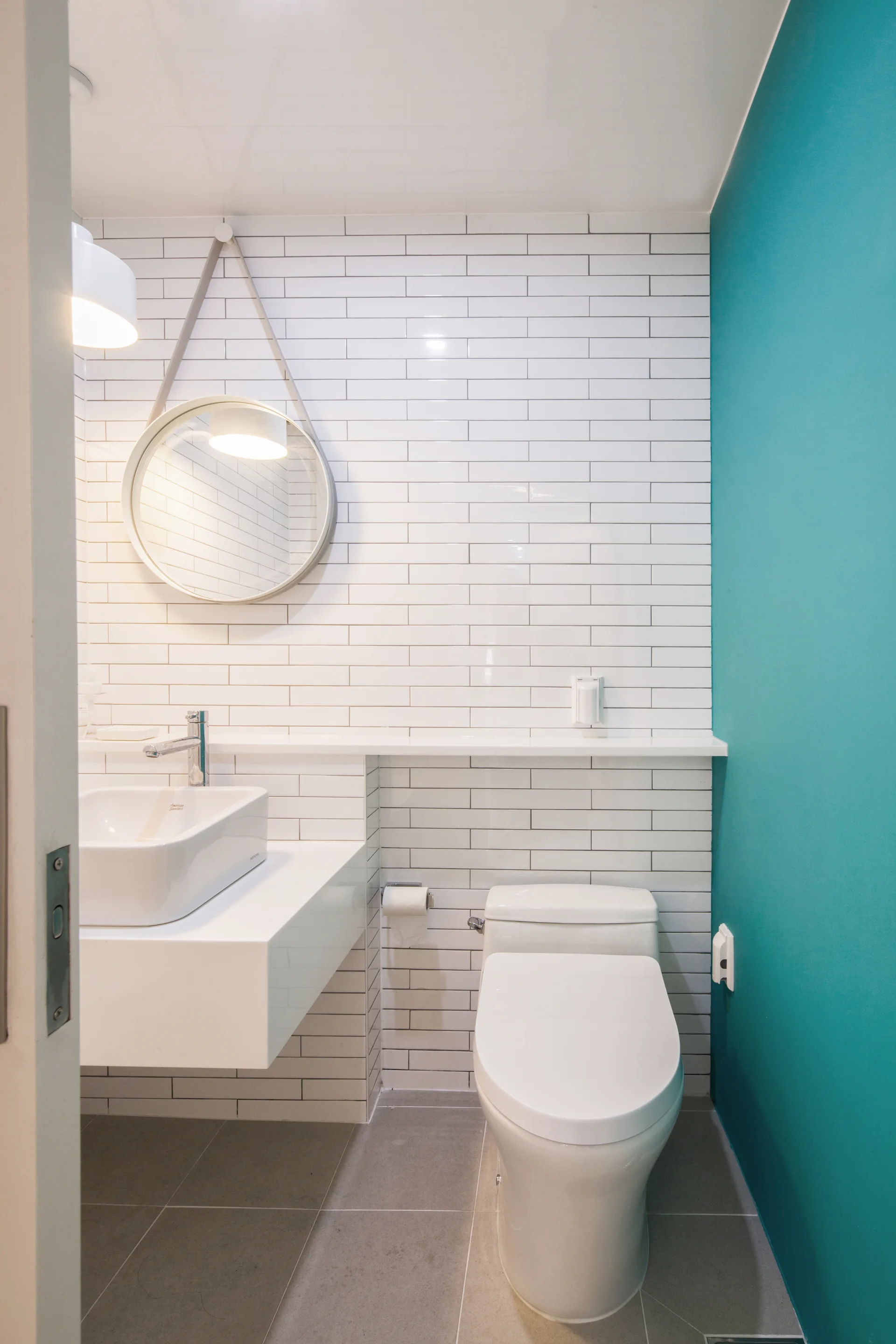 Modern bathroom featuring a white floating vanity, round strap mirror, white subway tiled wall, toilet, and vibrant teal accent wall.