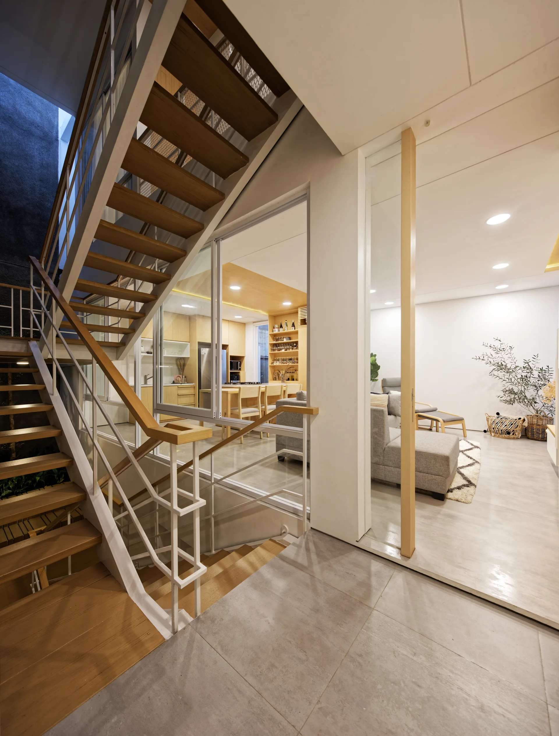 A modern wood-tread staircase with white metal railings is visible behind a glass partition wall looking into a neutral-toned living room and kitchen.