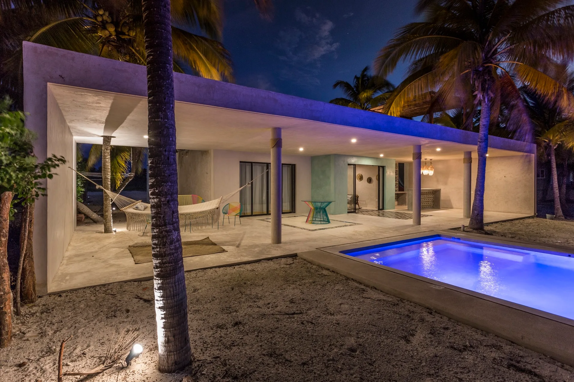 A nighttime view of a covered concrete patio featuring a glowing swimming pool, a white hammock, and colorful wire-frame outdoor furniture.