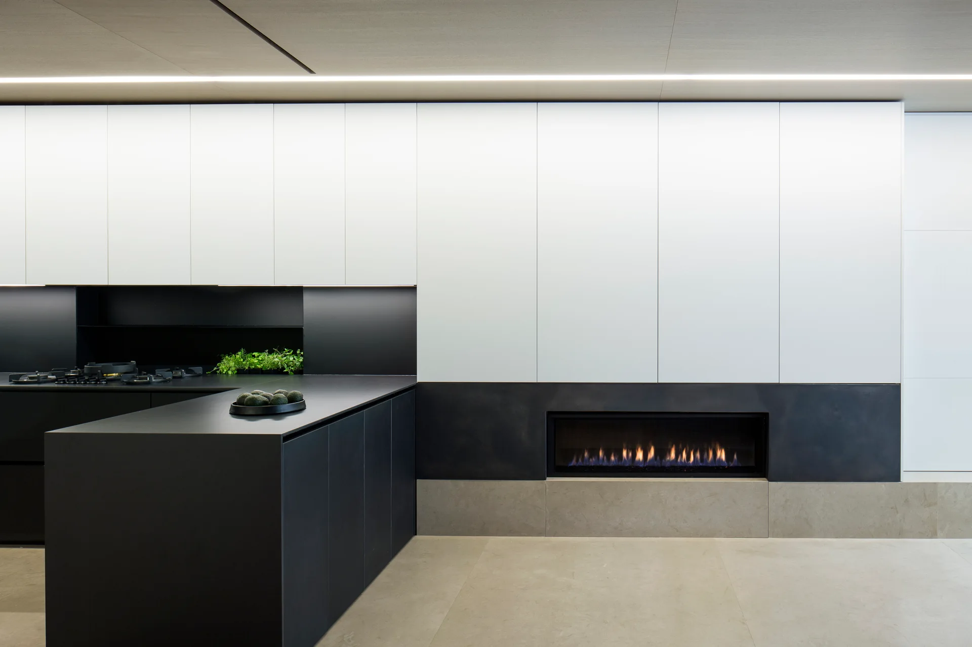 A minimalist kitchen featuring a matte black island, handleless white cabinetry, and an integrated linear fireplace built into the wall.