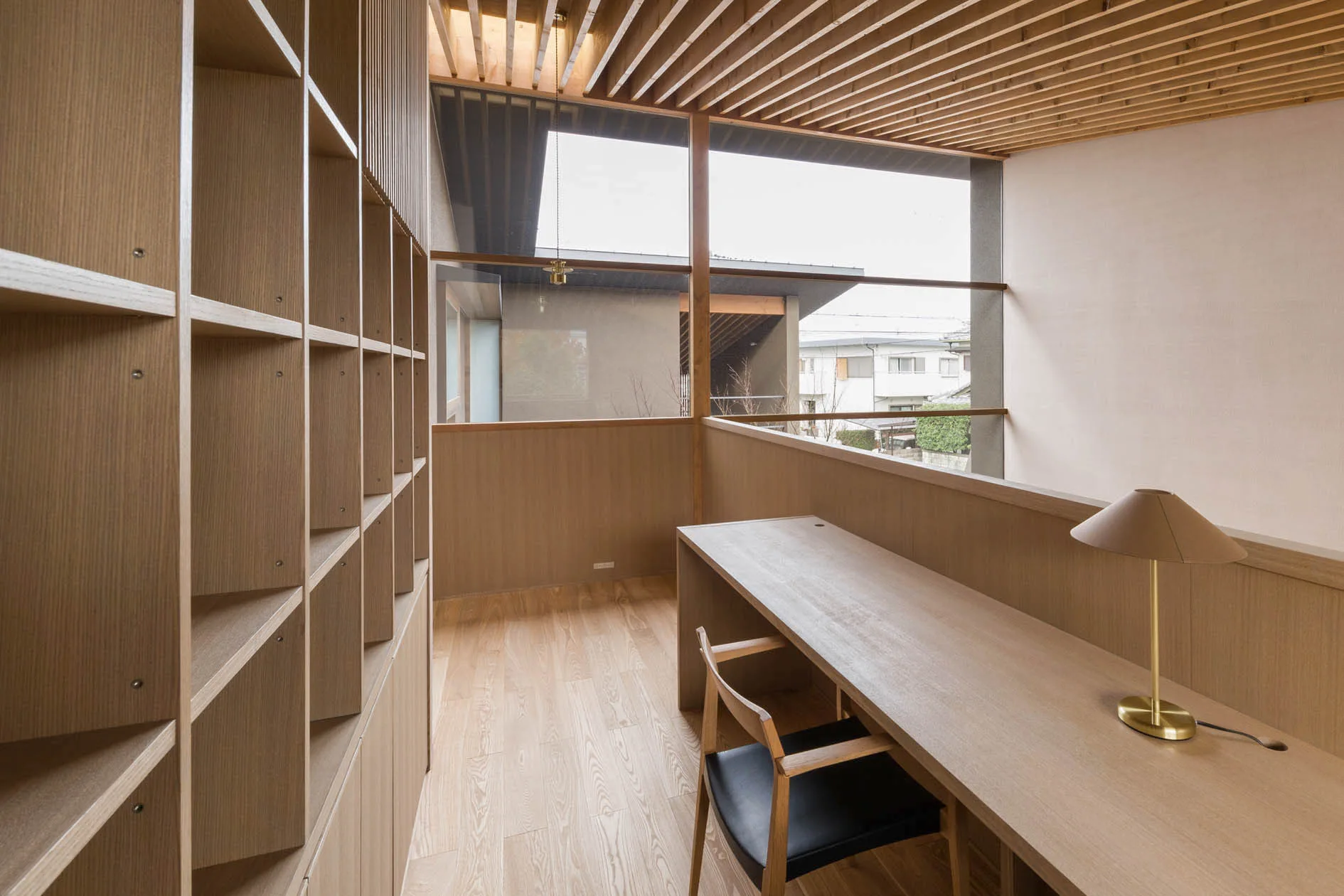 A minimalist wooden home office features a long desk, a matching chair, built-in bookshelves, and a slatted wood ceiling.