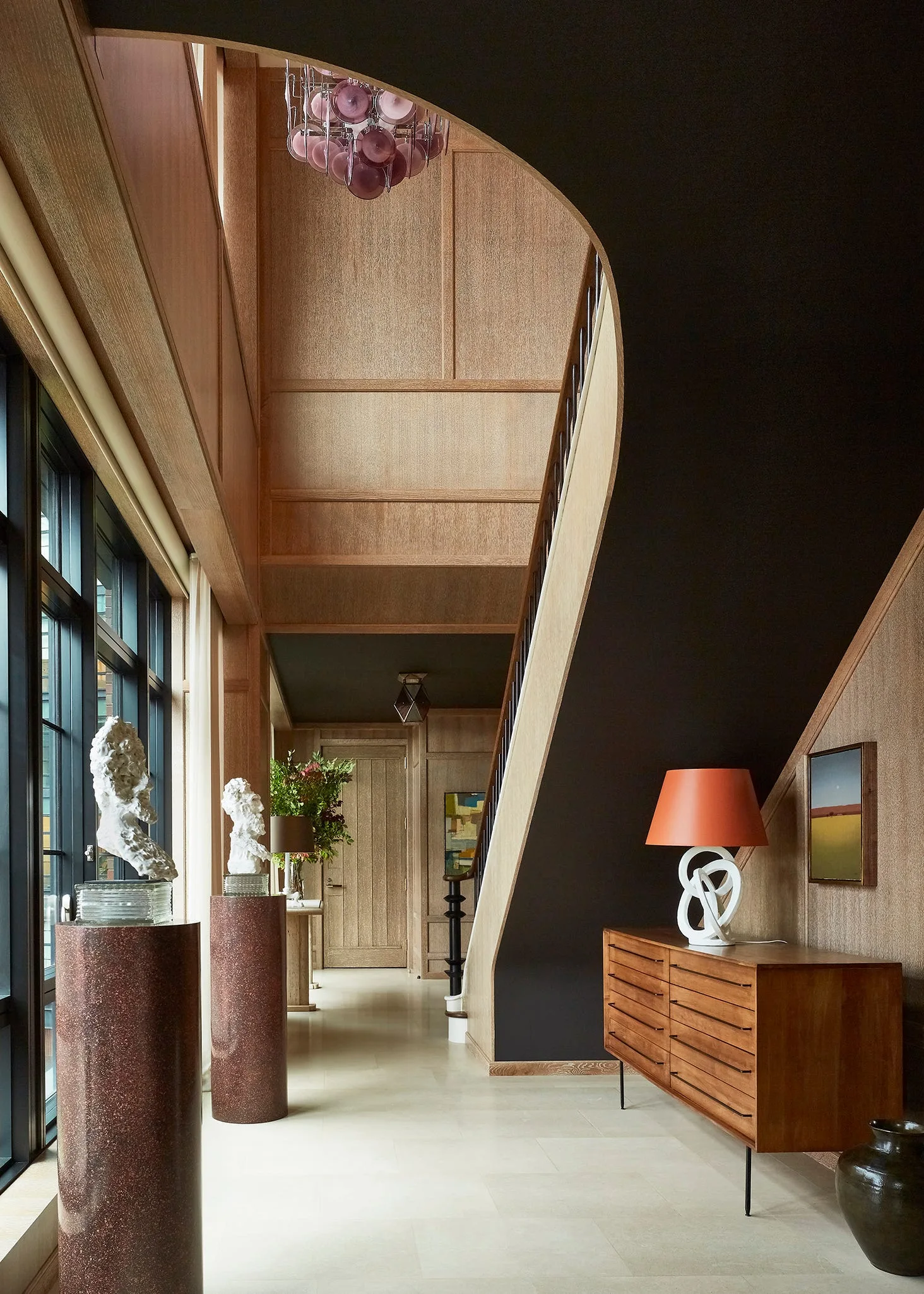 A foyer featuring wood-paneled walls, a curved staircase painted dark charcoal, a wooden console table with an orange lamp, and stone pedestals.